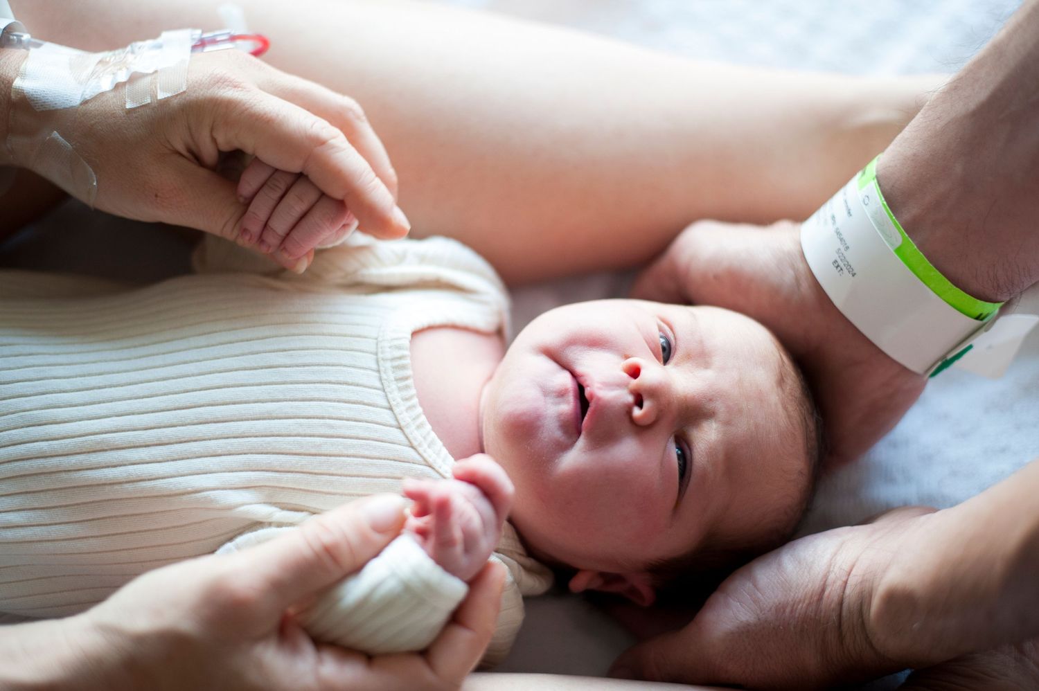 Newborn baby in hospital looks up between parents' hands.