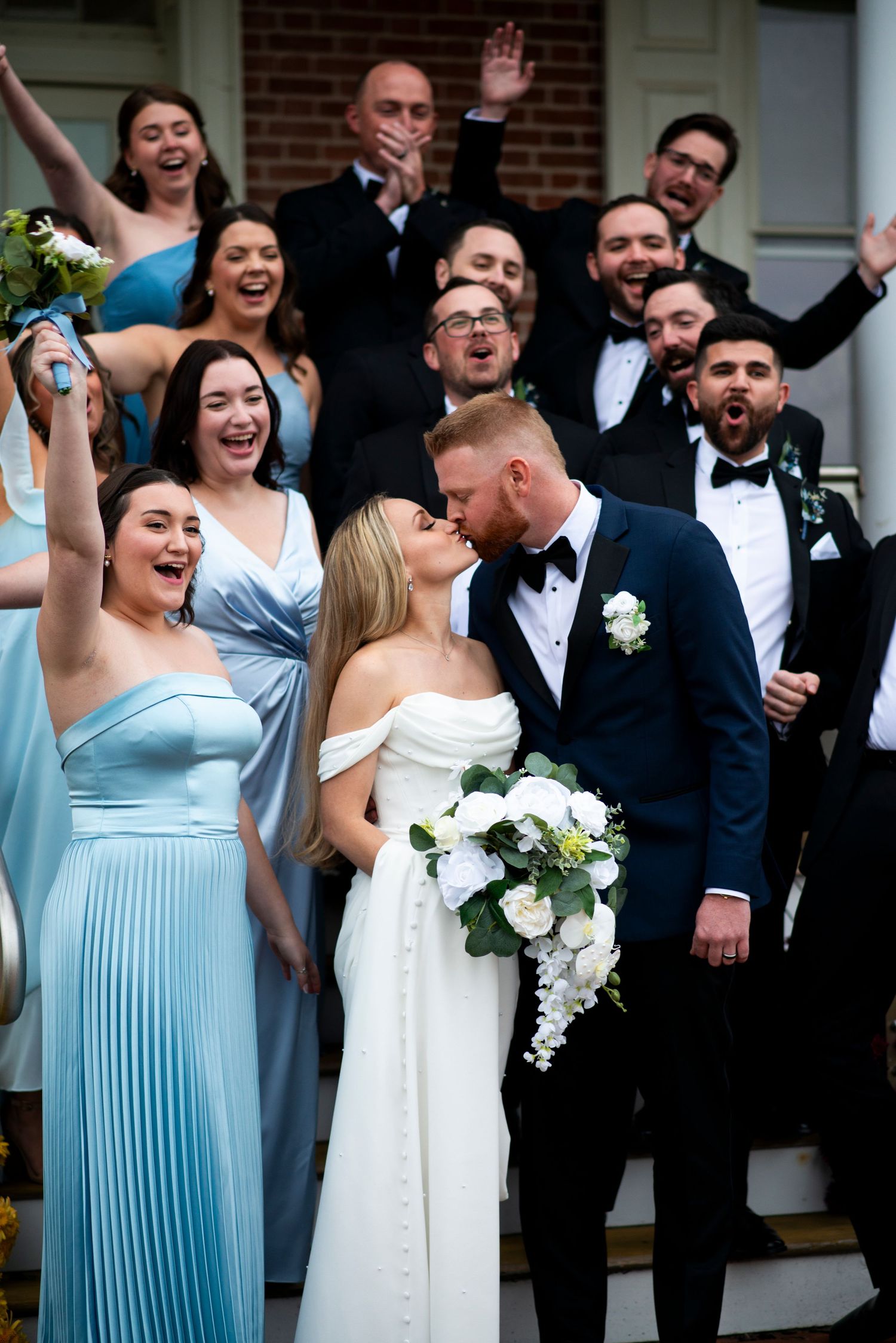 Bridal party in blue dresses and black suits cheers while bride and groom kiss at Lazaretto Ballroom in Pennsylvania.