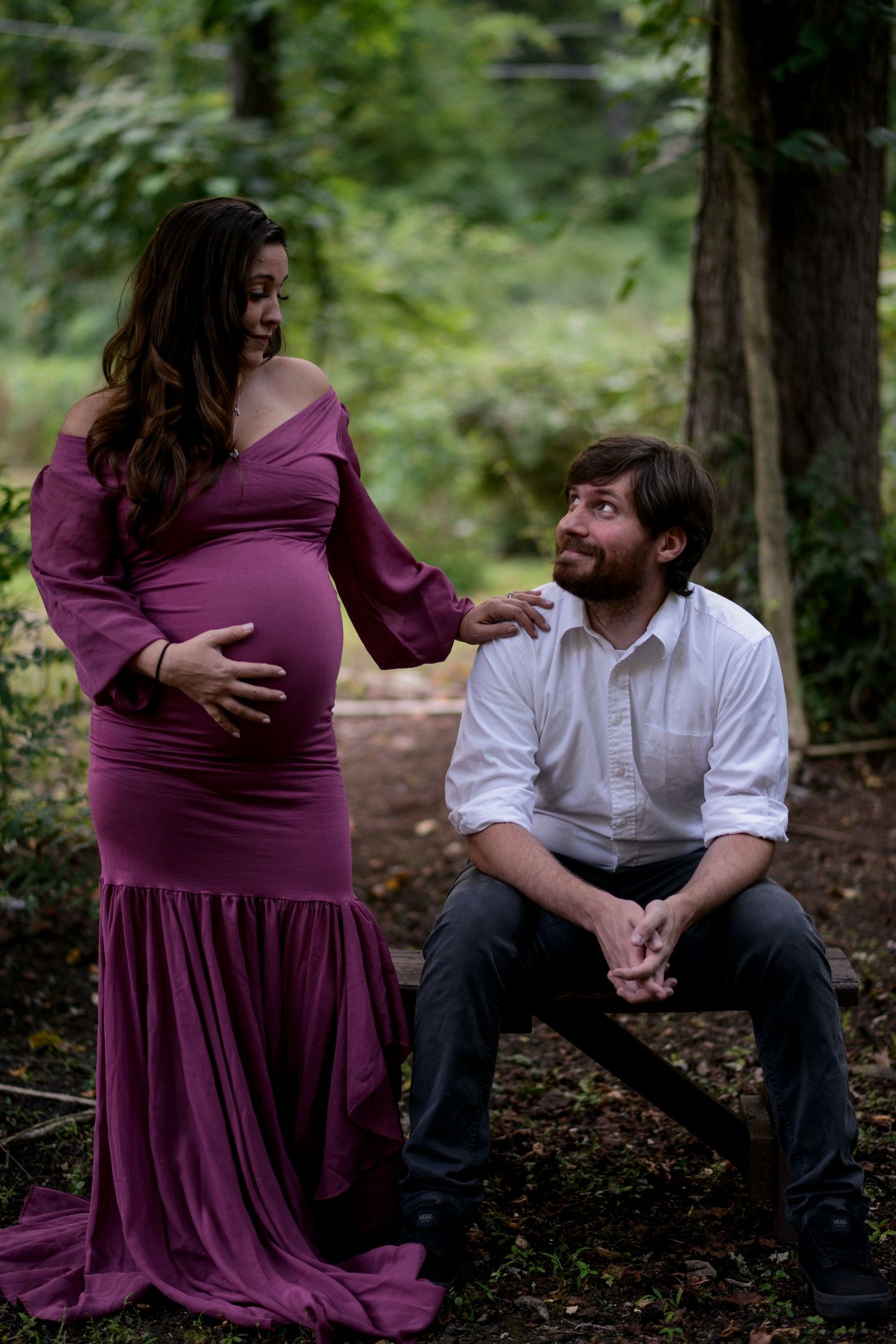 Pregnant woman in a dark mauve dress stands next to her seated husband with hand on her belly. They smile at each other.