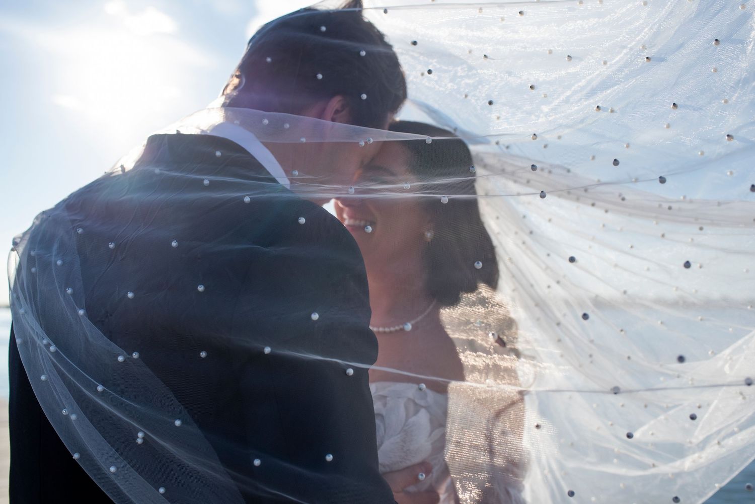 Bride and groom smile under the bride's veil as sunlight shines through.