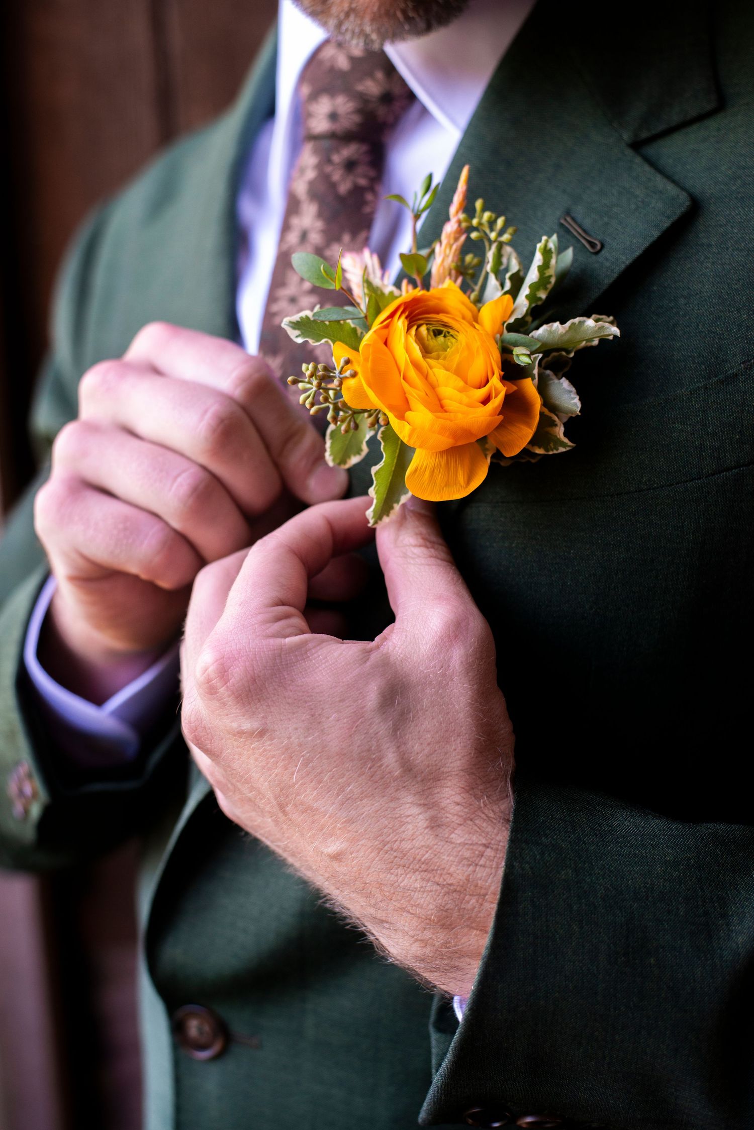Close up of groom in green jacket adjusting boutonniere with orange flower before his wedding ceremony at Bartram's Garden in Pennsylvania.