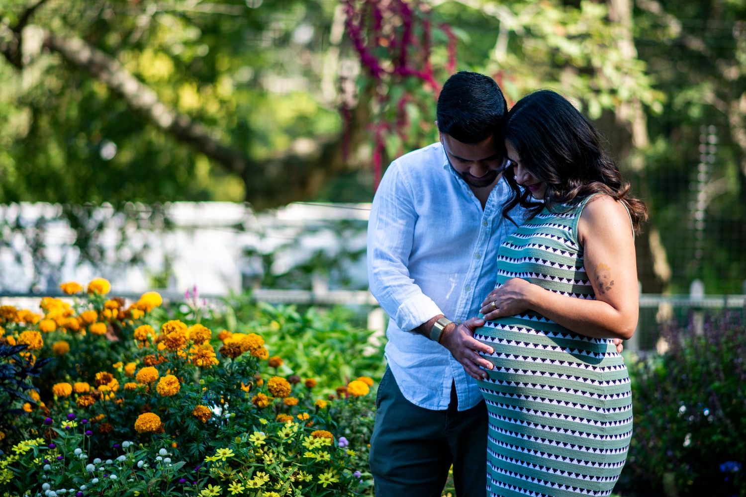 Pregnant couple hold belly and look down with eyes closed in front of orange marigolds at Deep Cut Gardens in New Jersey.