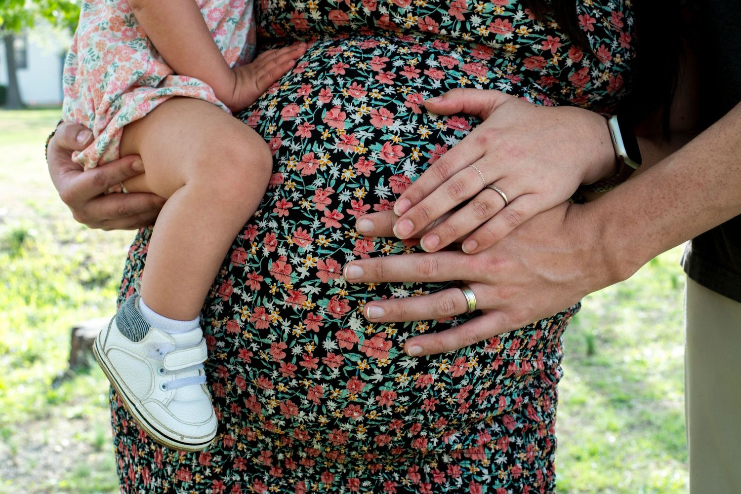 Closeup of mom and dad's hands on pregnant belly while mom is holding toddler in her arms.
