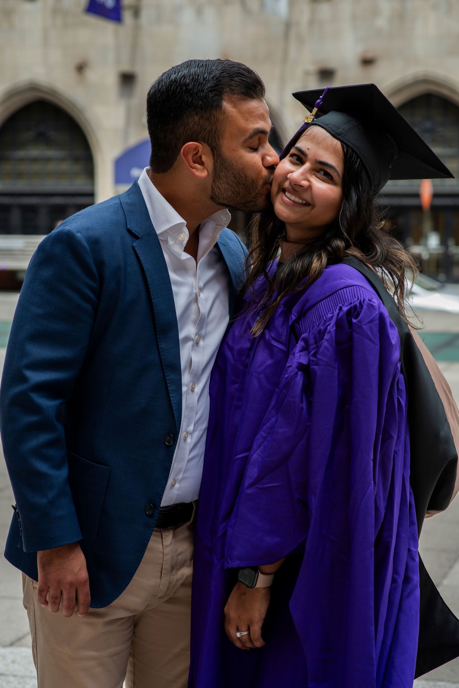 NYU Stern business school graduate smiles while her husband kisses her cheek on graduation day in New York City.