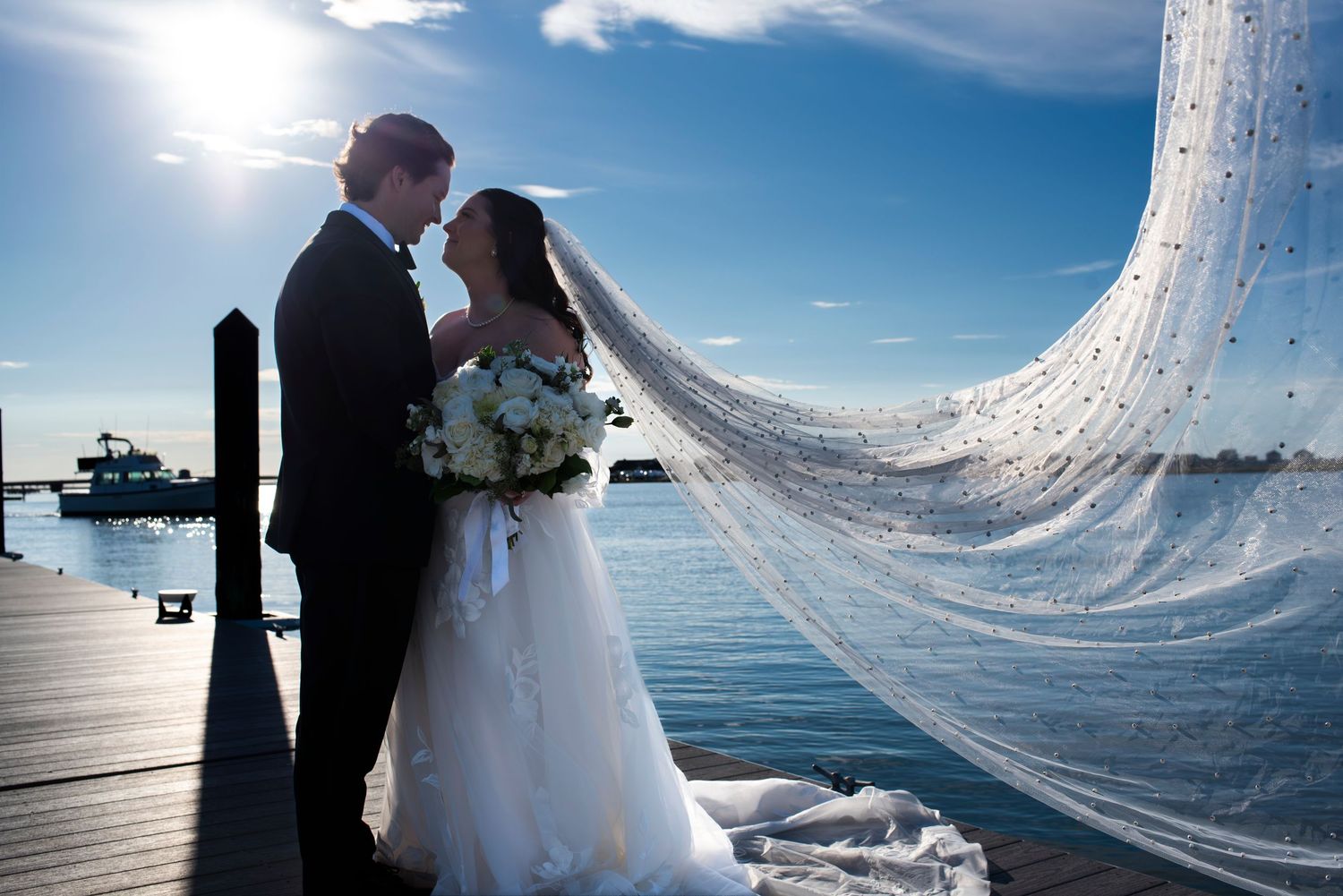 Bride and groom smile at each other in front of bright blue sky on the dock at Stone Harbor Yacht club. Bride's veil flows out behind her.