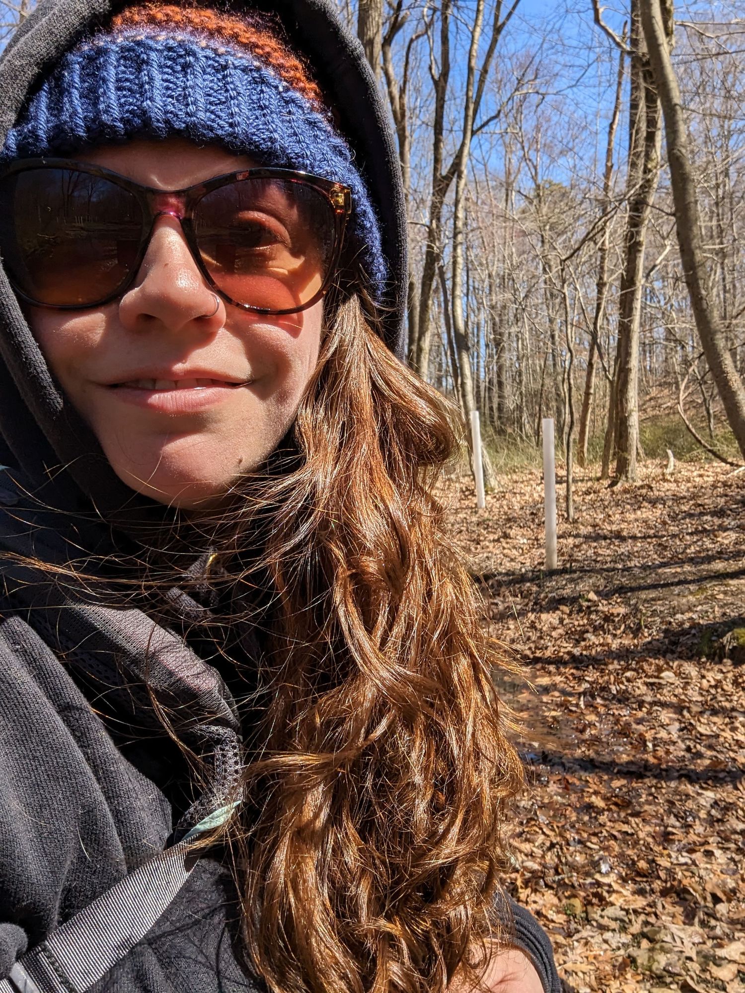 Selfie of woman with long, wavy hair and sunglasses hiking in the woods.