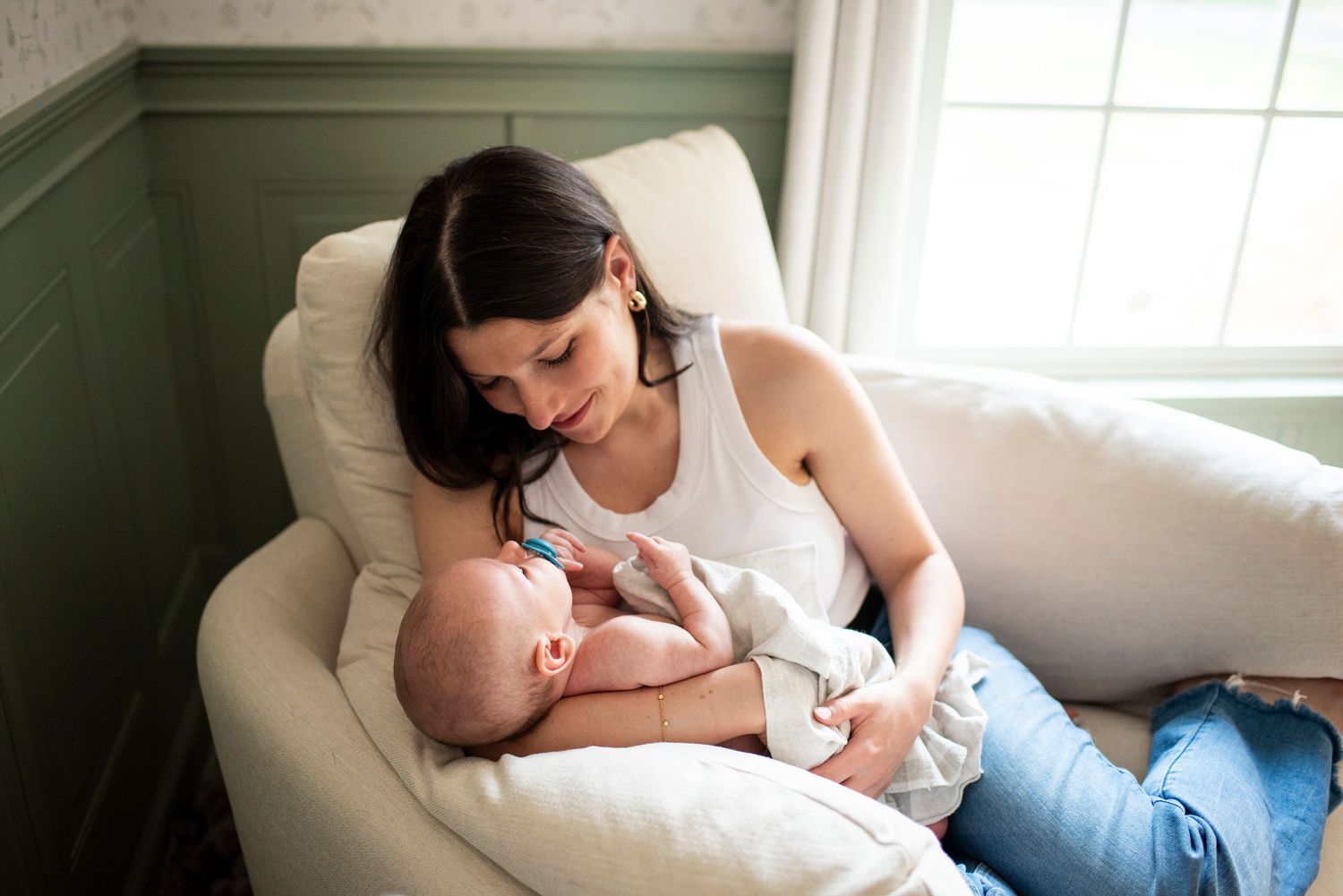 New mother smiles down at her newborn son in the window light of his nursery.