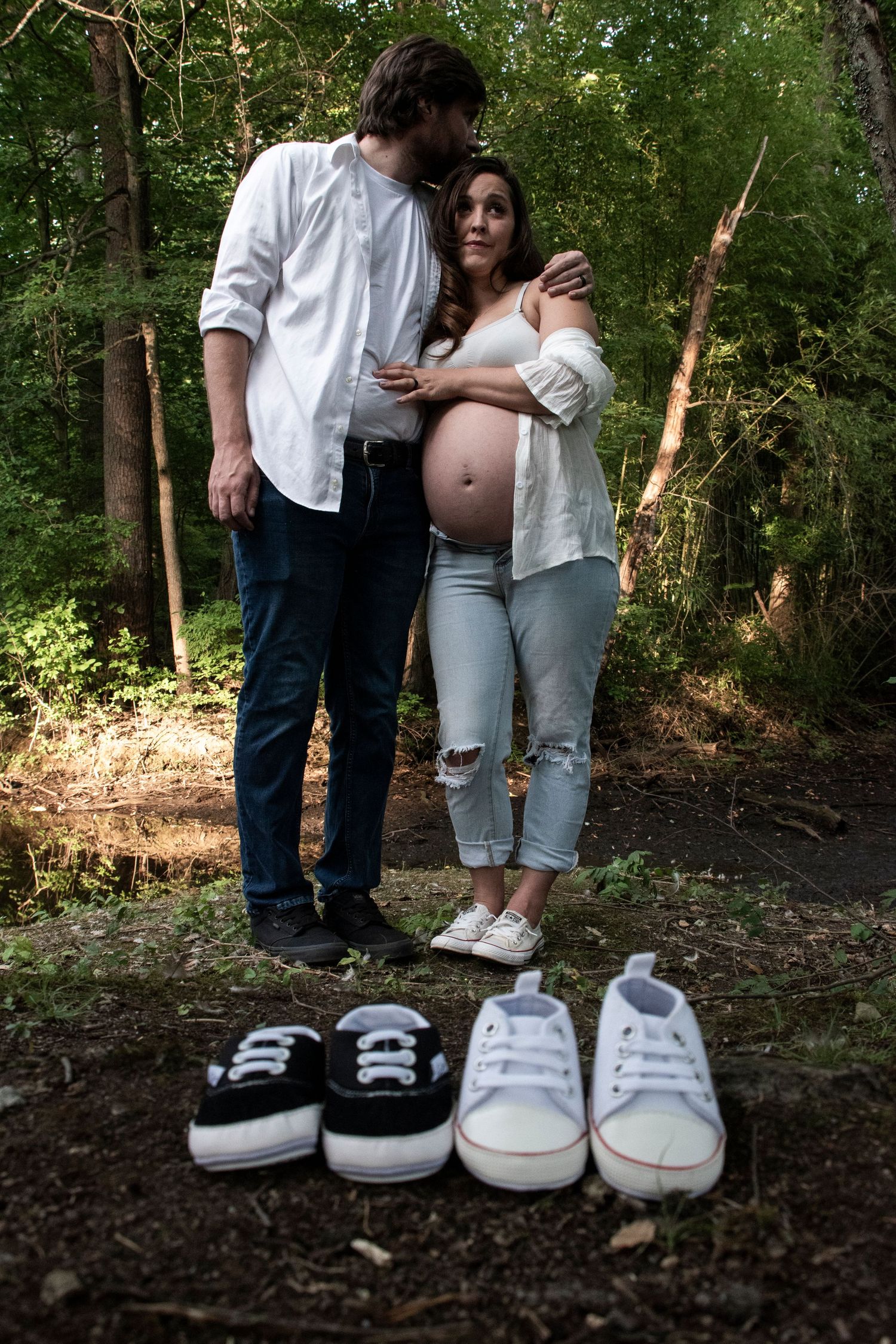 Man kisses his pregnant wife on the bank of a creek in New Jersey. Two tiny pairs of shoes are in the foreground.