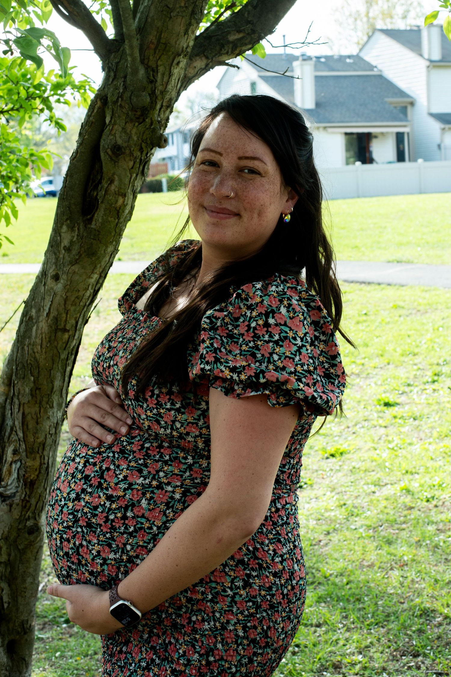 Pregnant mother smiles for the camera holding her belly under a tree.