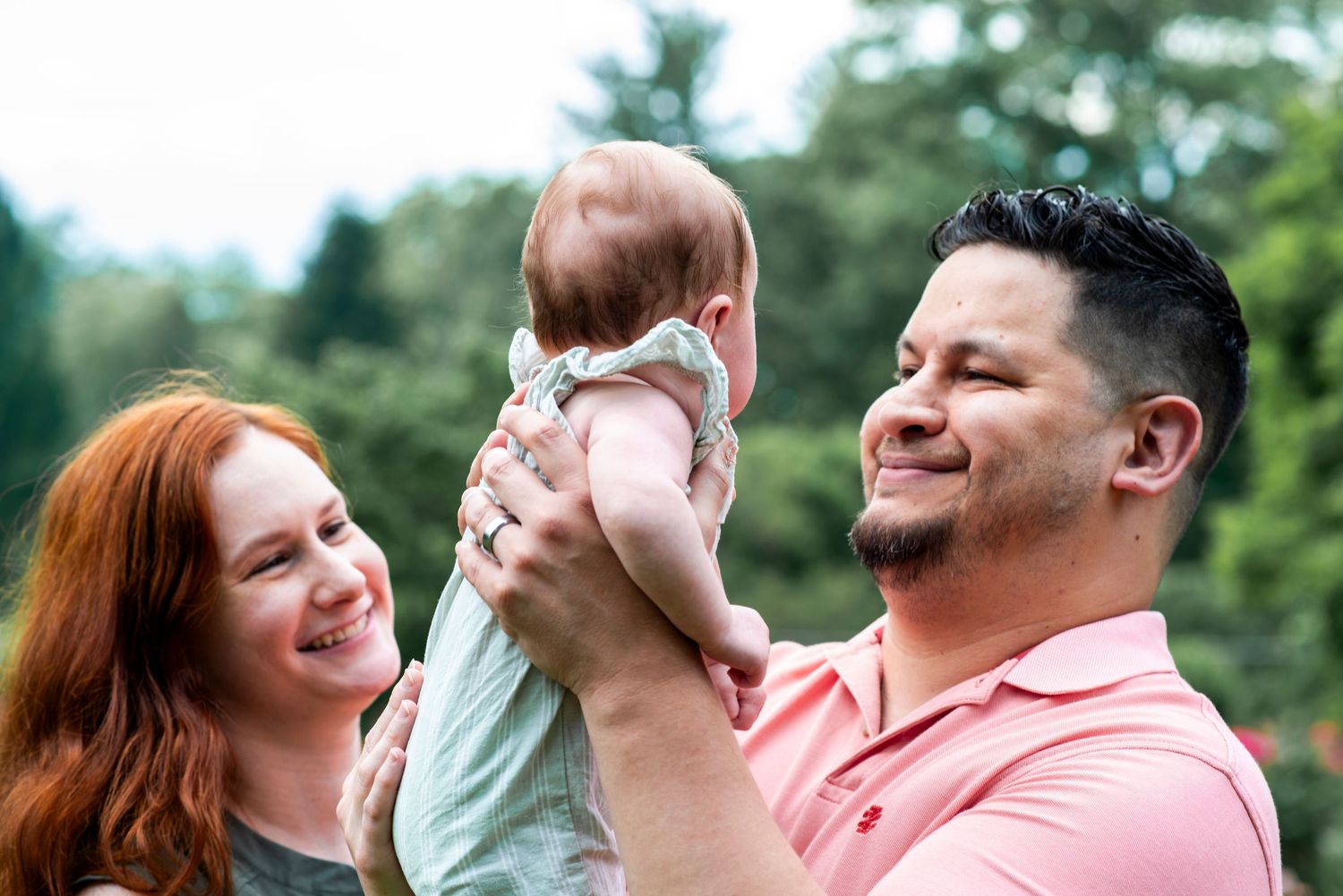 Outdoor portrait of parents smiling as they hold infant daughter up to look at her,