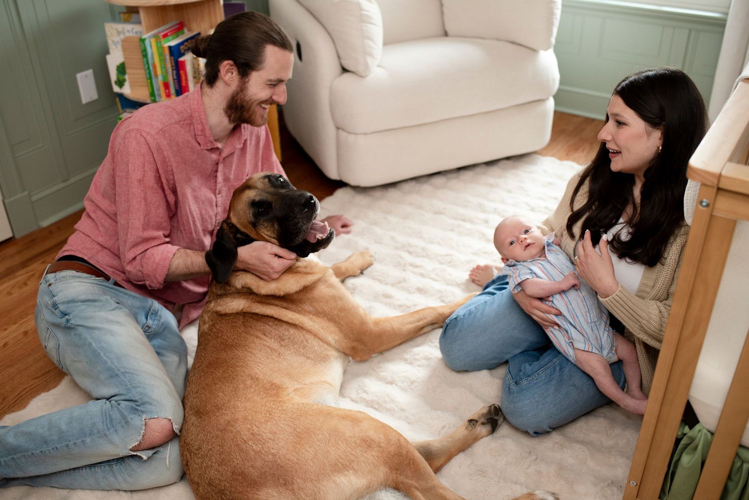 New parents and newborn baby with their great dane smile at each other sitting on the nursery floor in their home in New Jersey.