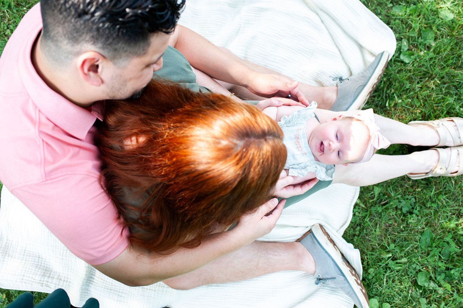 Family of three portrait in the grass from above as baby smiles up from mom's lap.