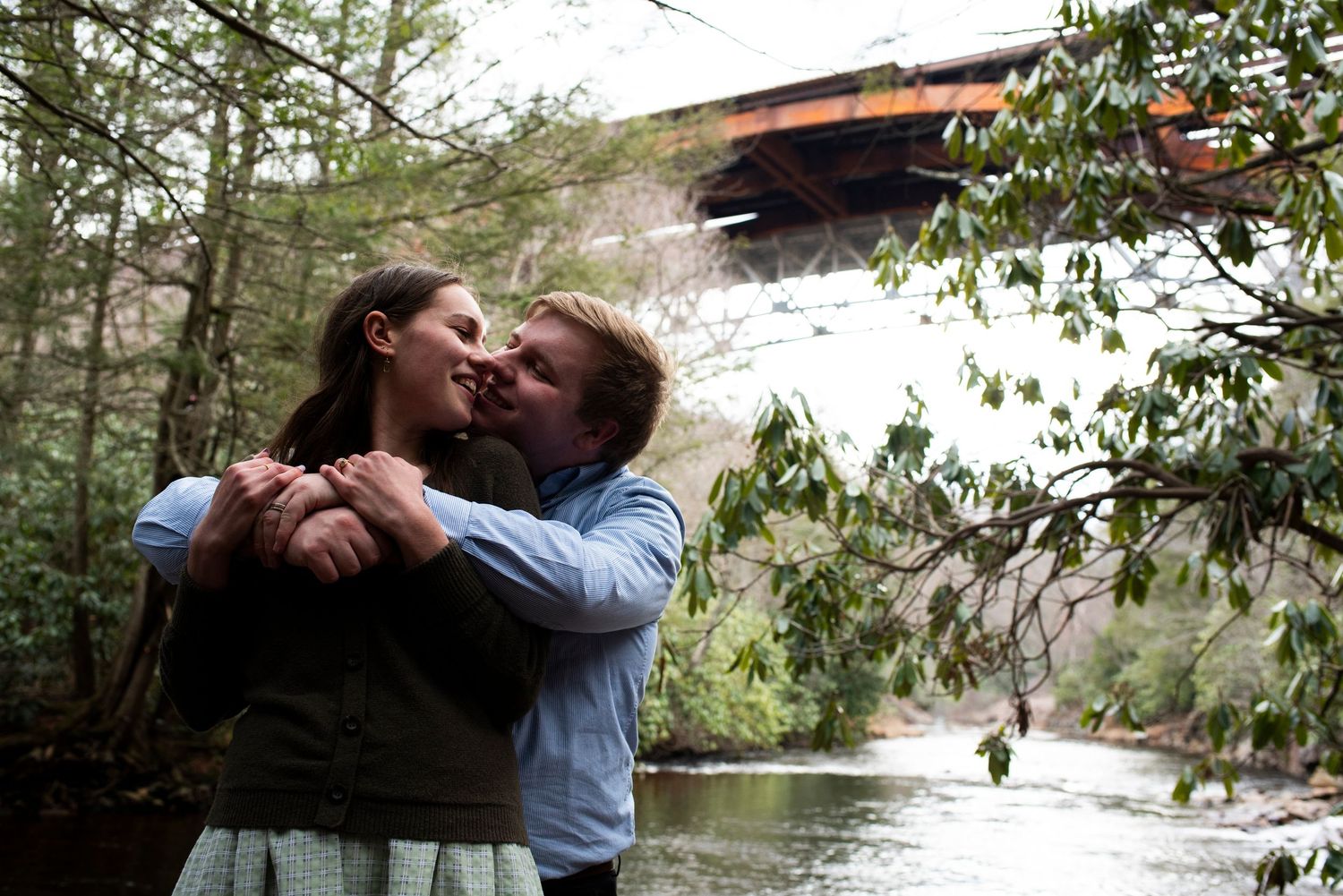 Engaged couple kisses with arms around each other with river and bridge in the background at Hawk Falls in Pennsylvania.