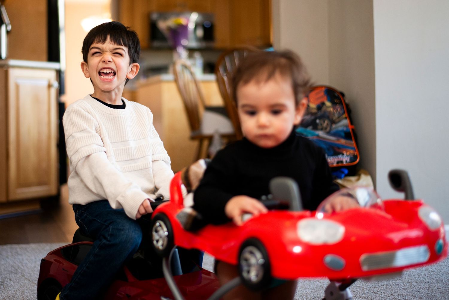 Brother and sister ride toy cars around their living room together.