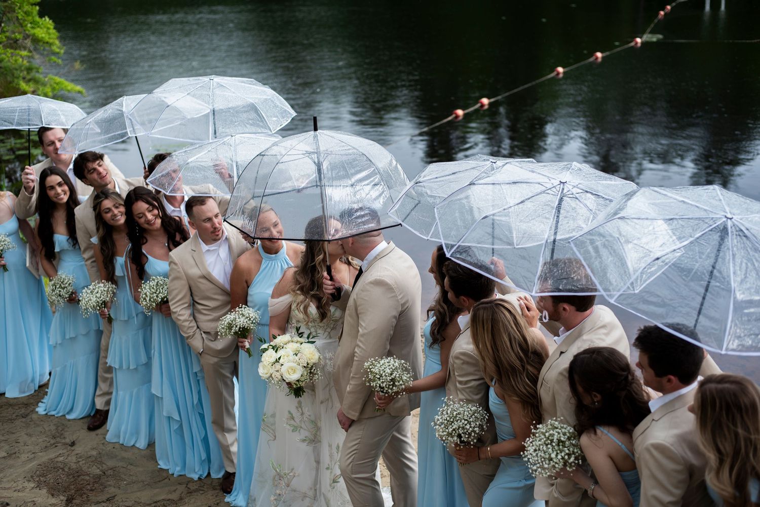 Bridal party looks on from under a line of umbrellas as bride and groom kiss on the beach at Camp Matollionequay in Medford, New Jersey.