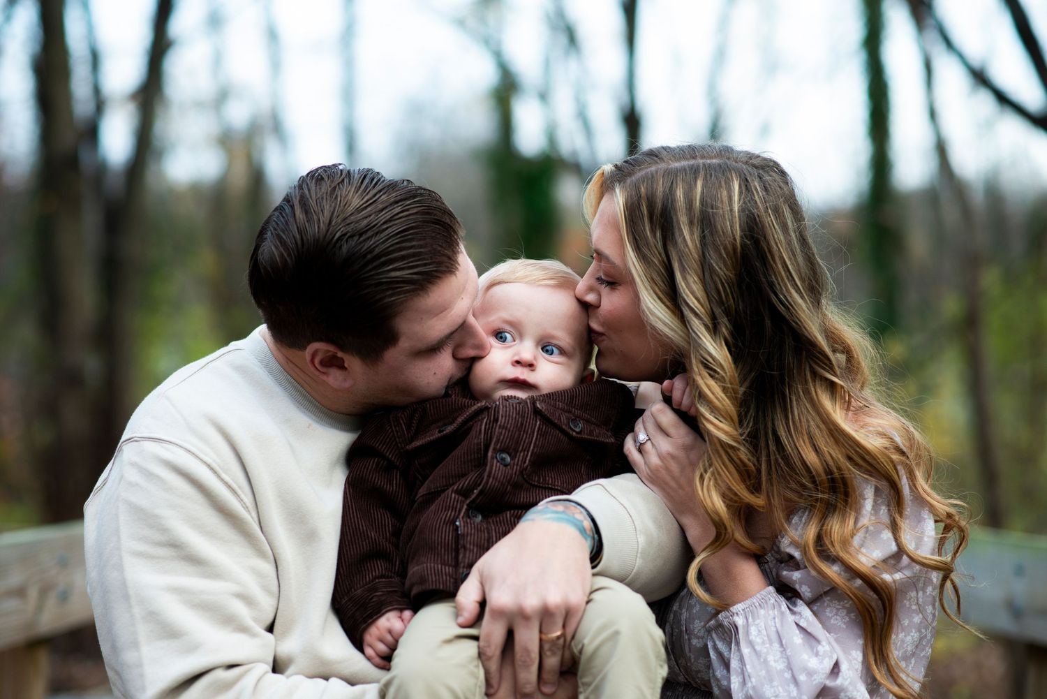 Parents squish toddler's cheeks with a kiss in outdoor family portrait.