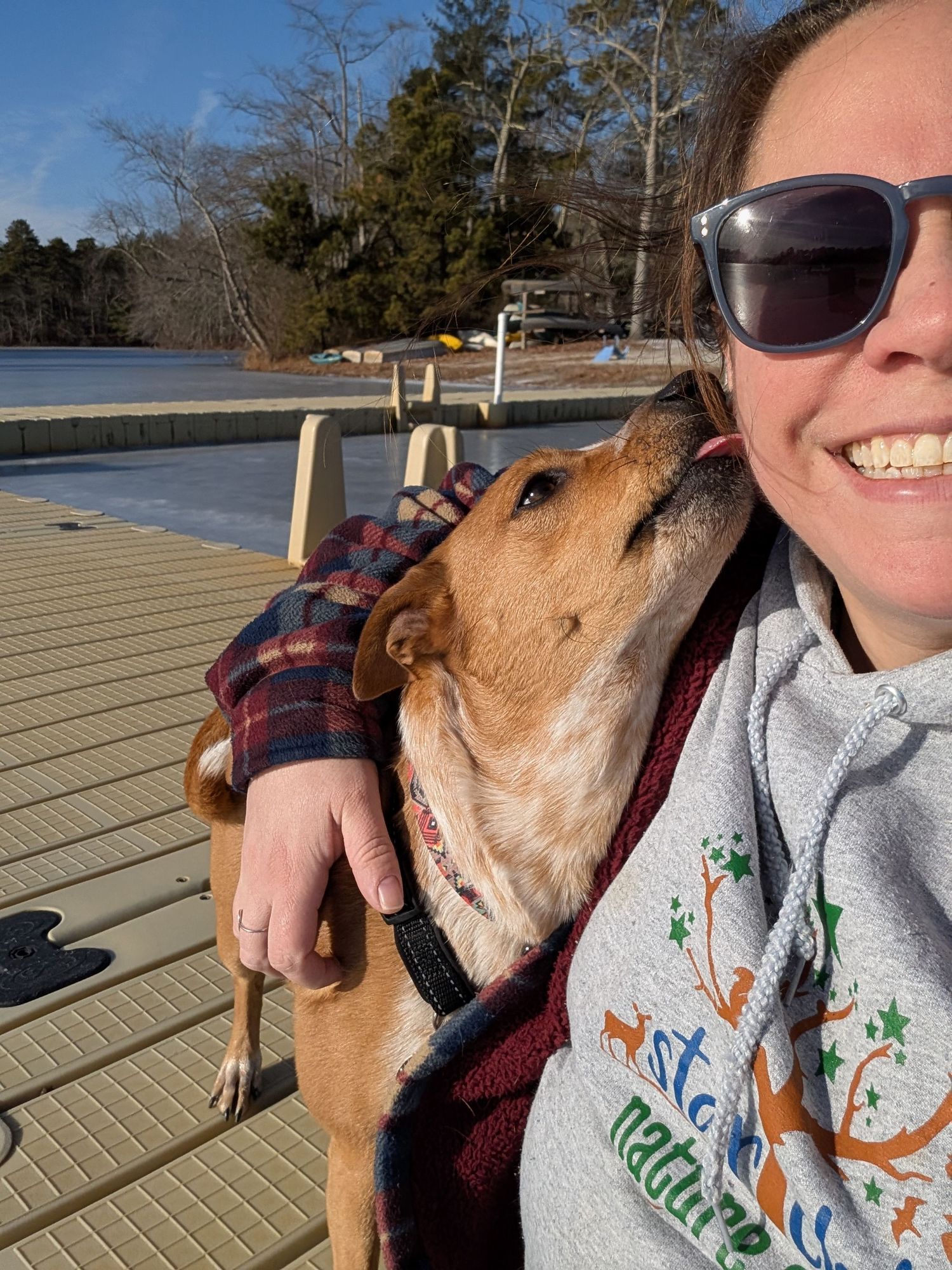 Selfie of smiling woman in sunglasses sitting on a dock while dog licks her cheek.