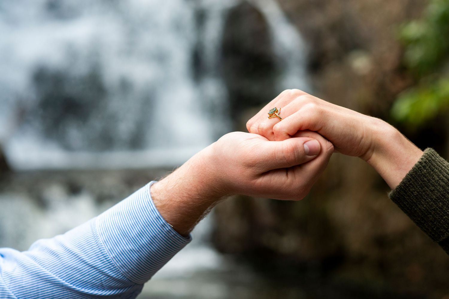 Engaged couple holds hands showing off engagement ring in front of waterfall in Hawk Falls, PA.