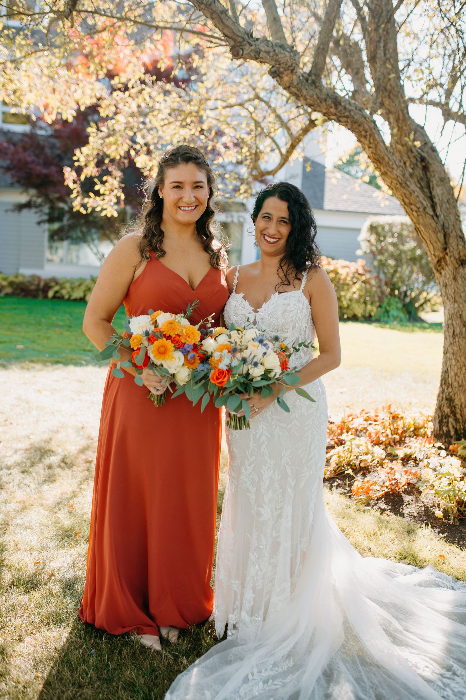 Two women pose under blooming tree holding orange and white bouquets.