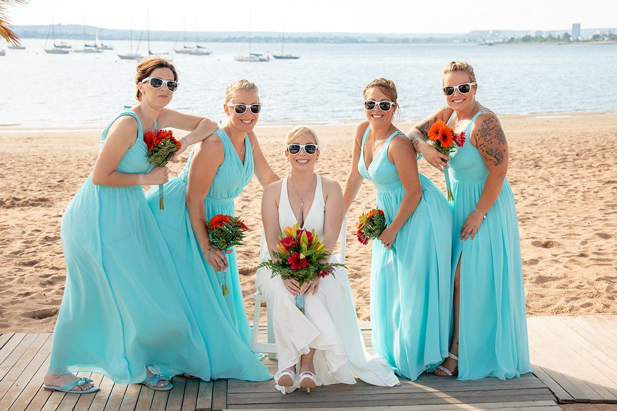 Bridal party in turquoise dresses poses together on a sunny beach with ocean views and tropical flowers.