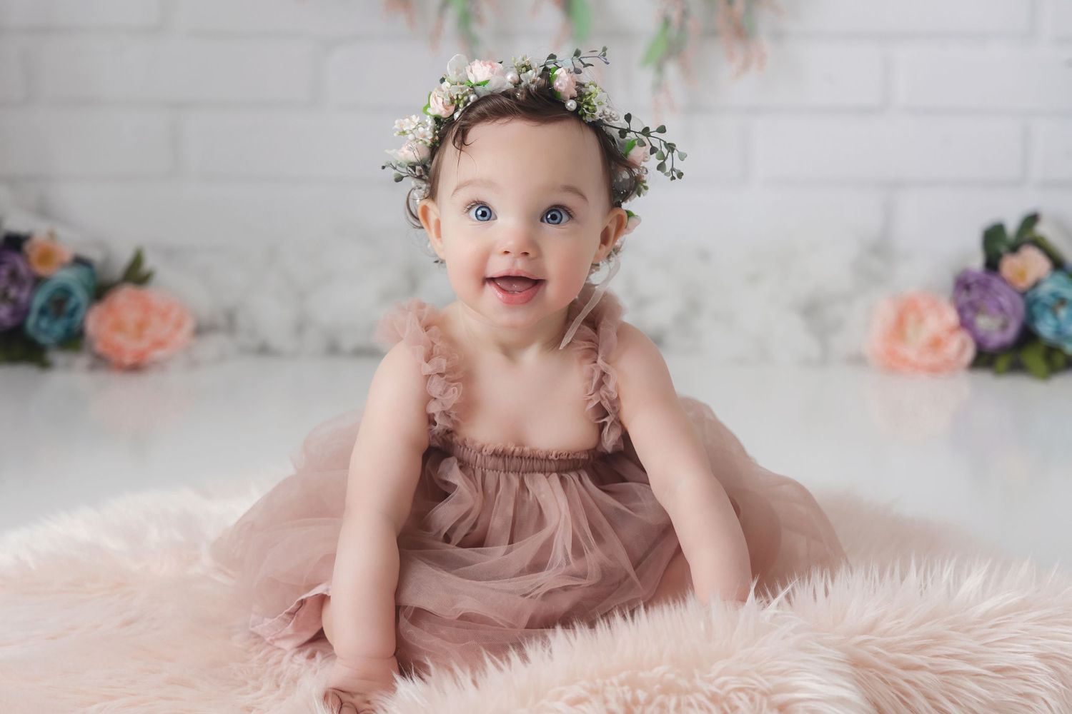 Baby in pink dress sits on fluffy white rug with floral crown and pastel decorations in background.