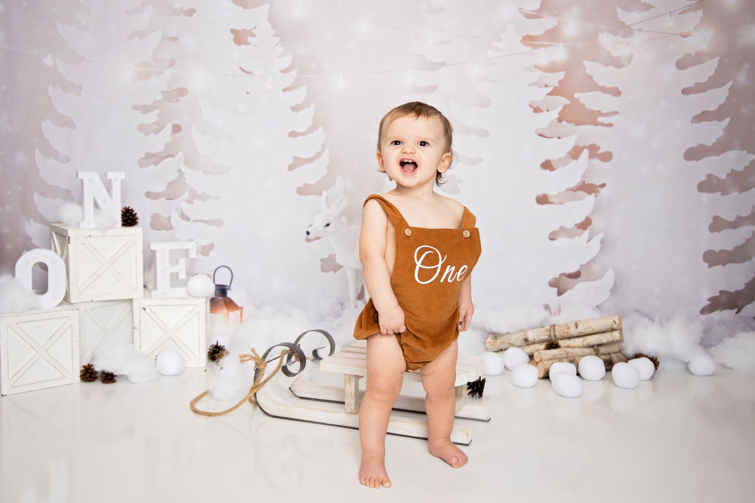 A baby wearing a brown bodysuit stands in a winter-themed photo setup with white trees and decorative elements in the background.