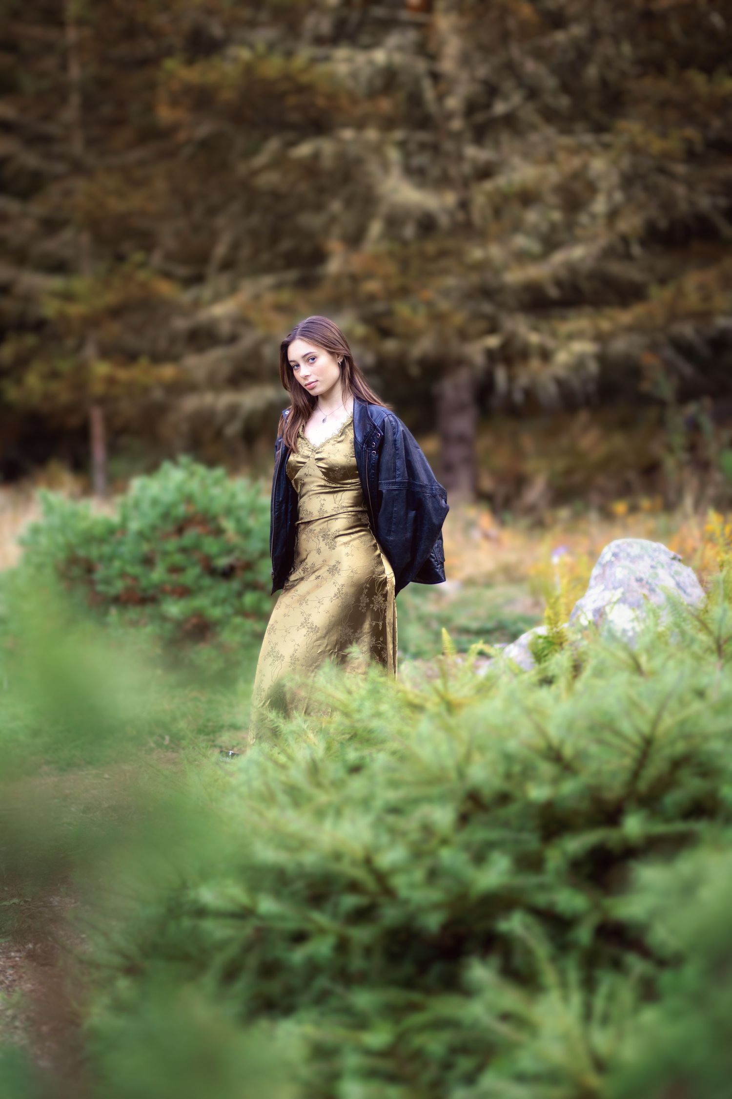 A person in a gold dress and navy jacket poses elegantly among green foliage in an outdoor garden setting.