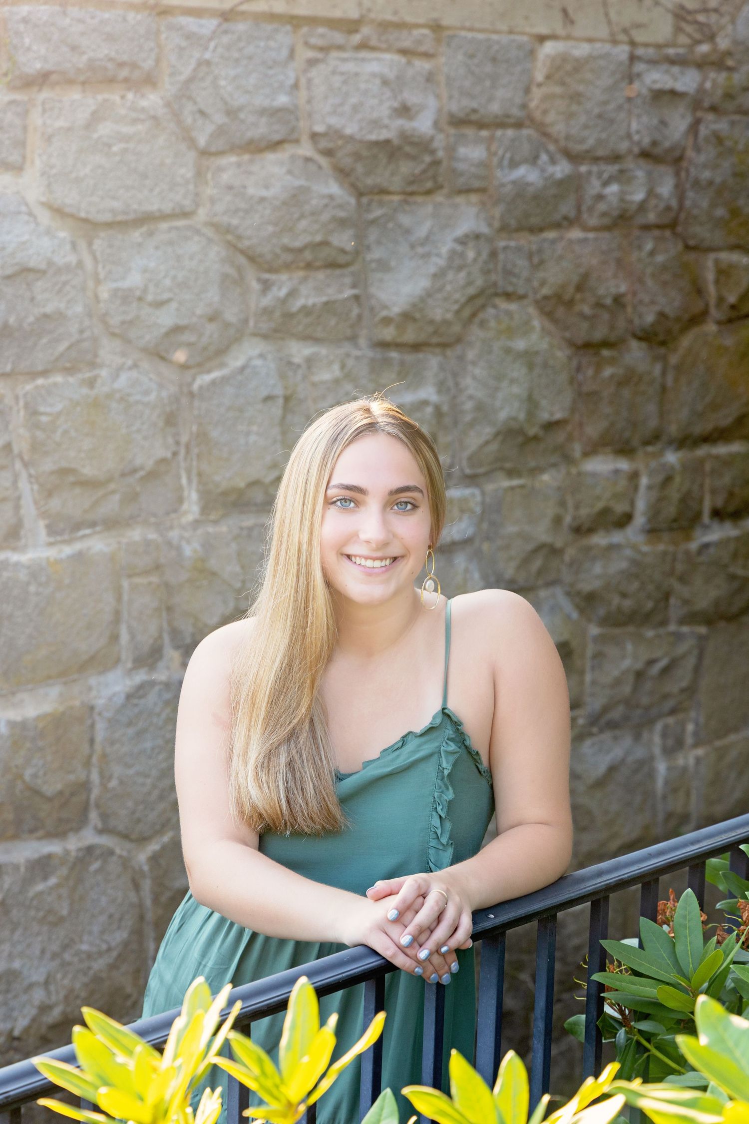 A smiling figure in a green dress leans against a railing with yellow lilies and a stone wall backdrop.