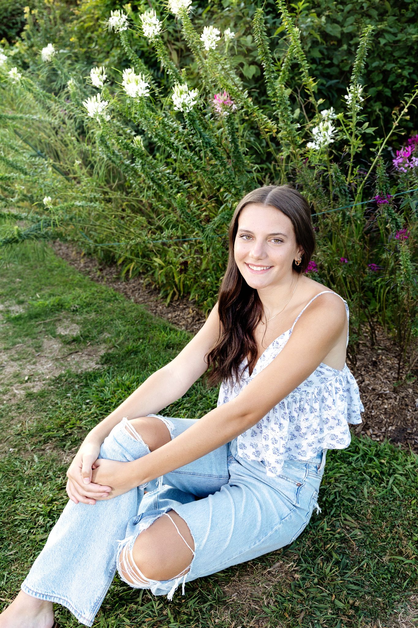 A person in a blue and white floral dress relaxes on grassy ground near blooming flowers and greenery.