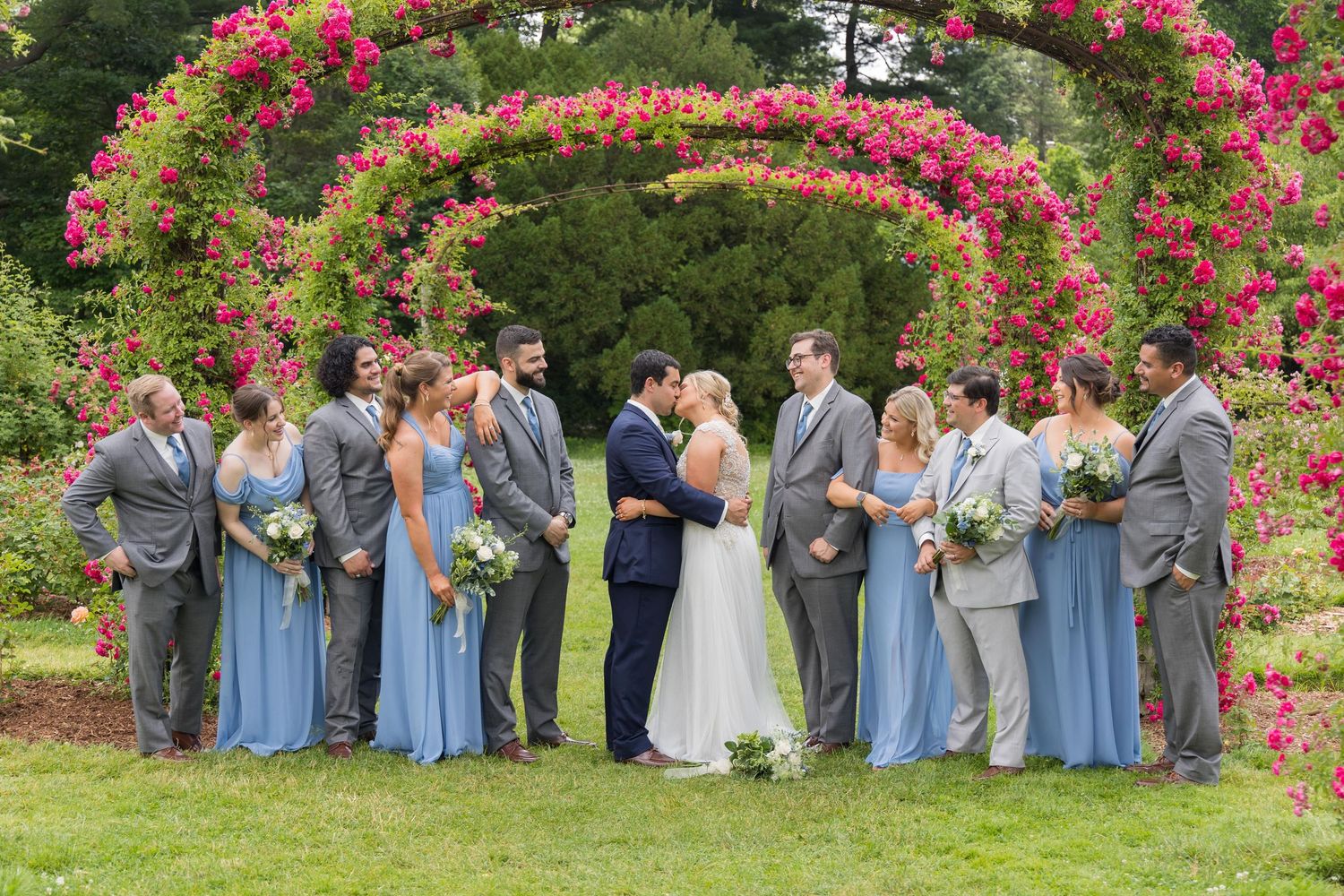 Wedding party poses under vibrant pink floral archway in garden setting wearing blue and gray attire.