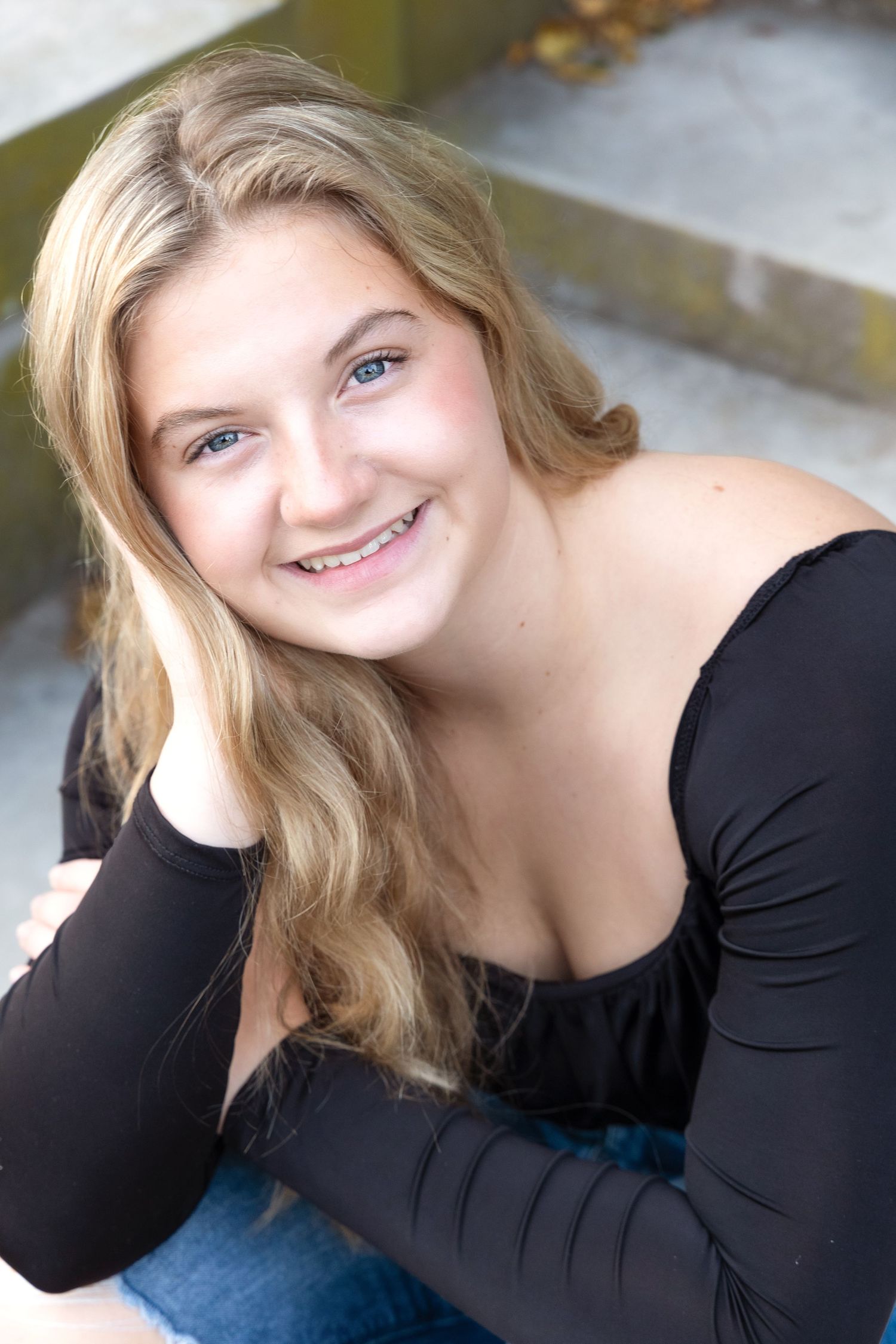 Portrait photo of someone with long blonde hair wearing a black long-sleeved top and blue jeans while sitting outdoors.