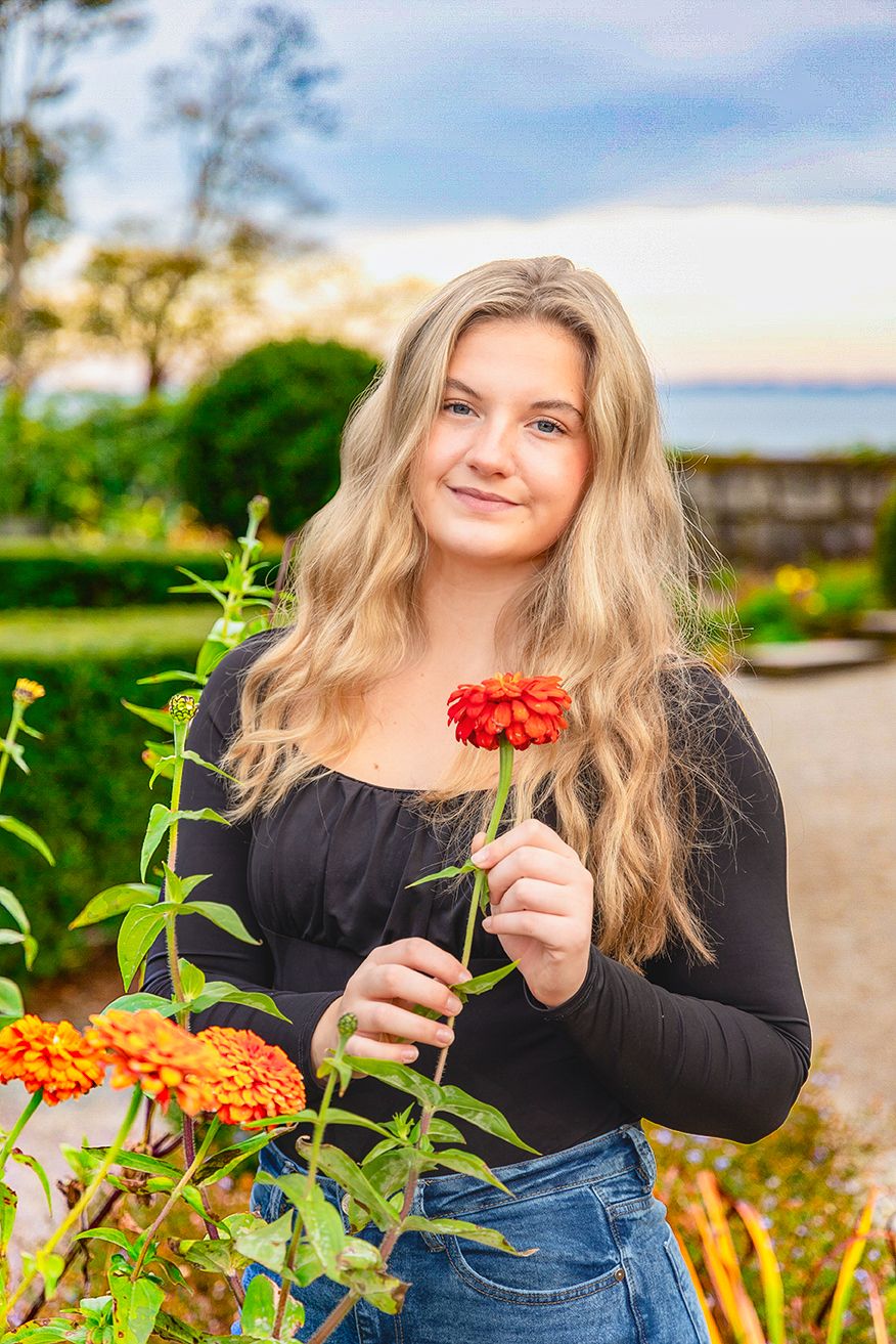 Person in black top and jeans poses with vibrant orange zinnia flowers in a sunny garden.