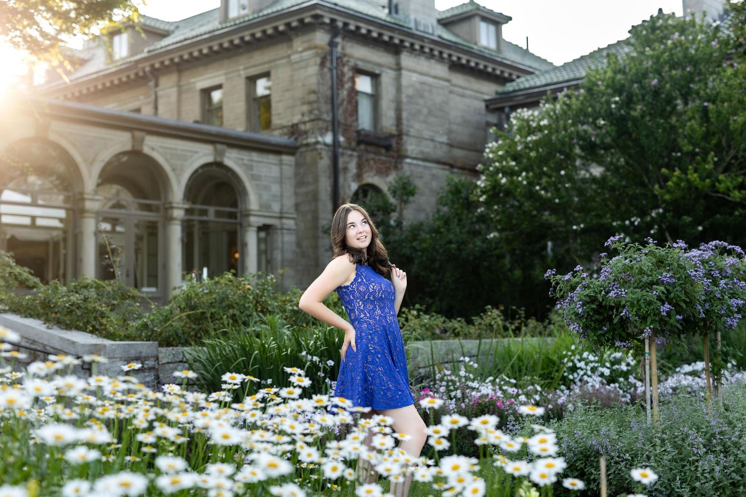 A person in a blue dress stands among blooming daisies in front of an elegant stone mansion at sunset.