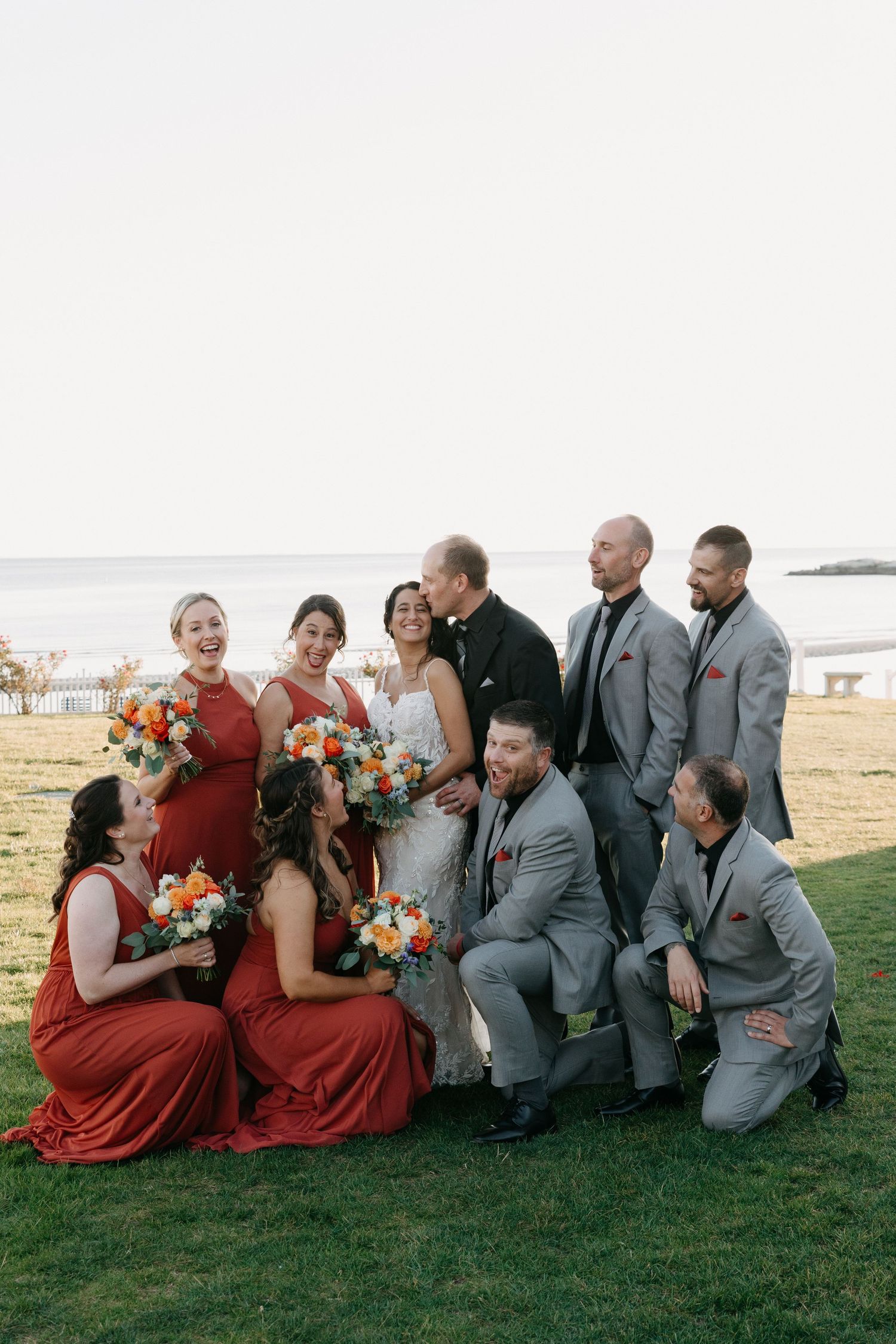 Wedding party gathered on grass by the ocean in red dresses and gray suits.