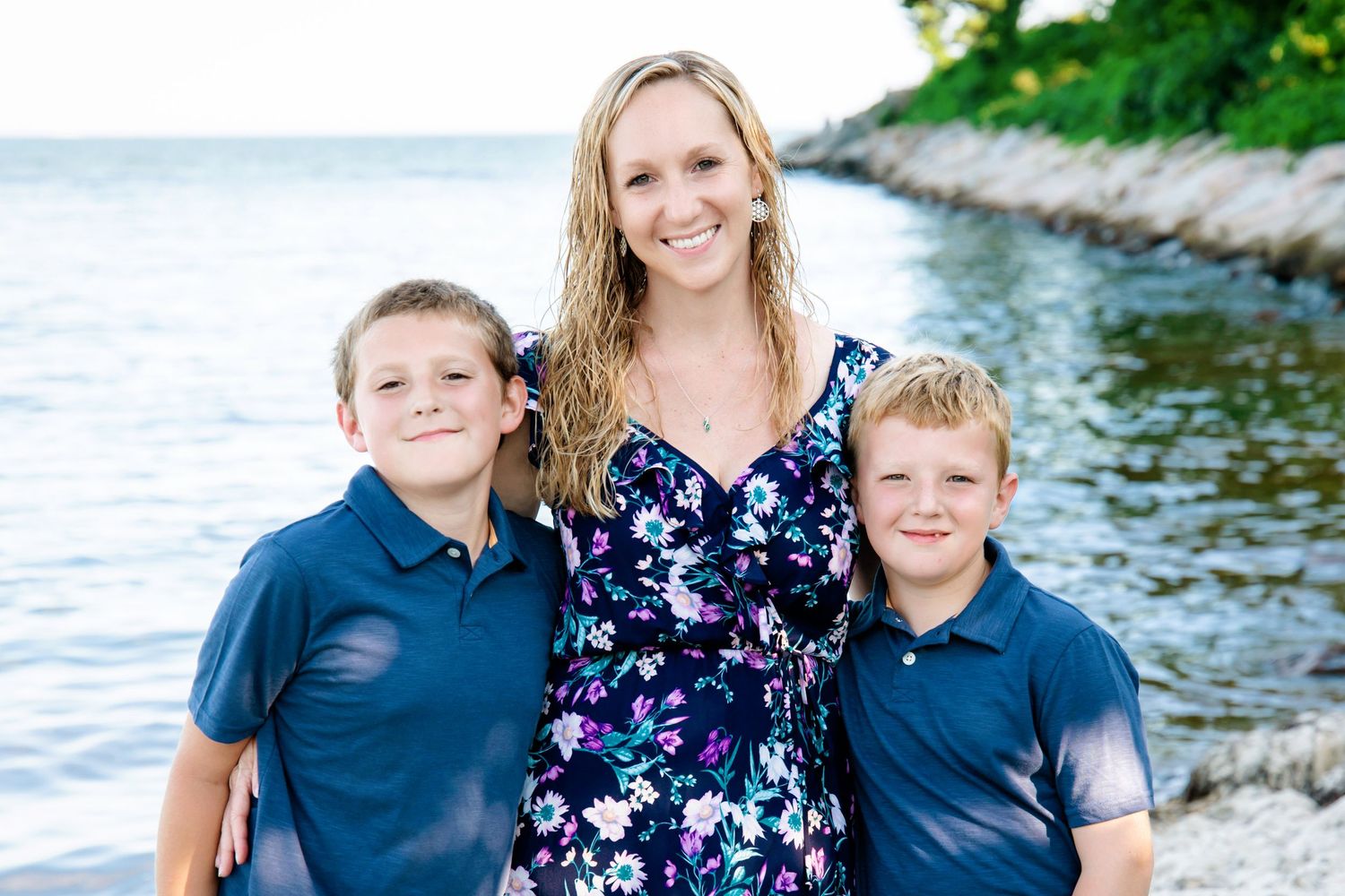 Family in coordinated blue outfits poses together along a rocky shoreline on a sunny summer day.