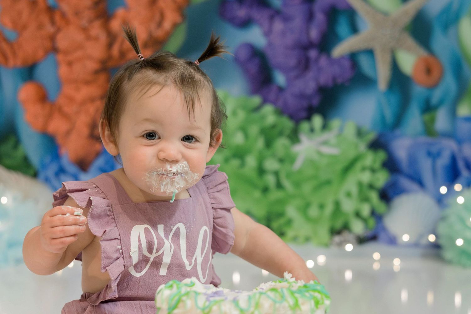 A baby in a pink dress sits on a colorful backdrop during a cake smash photoshoot celebration.