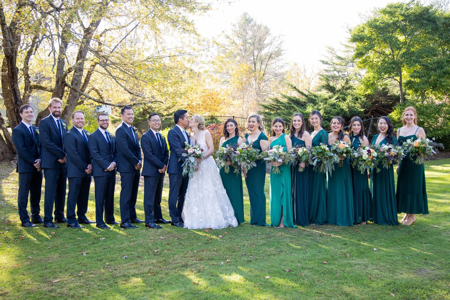 Large wedding party poses outdoors in navy blue and emerald green attire during autumn season.