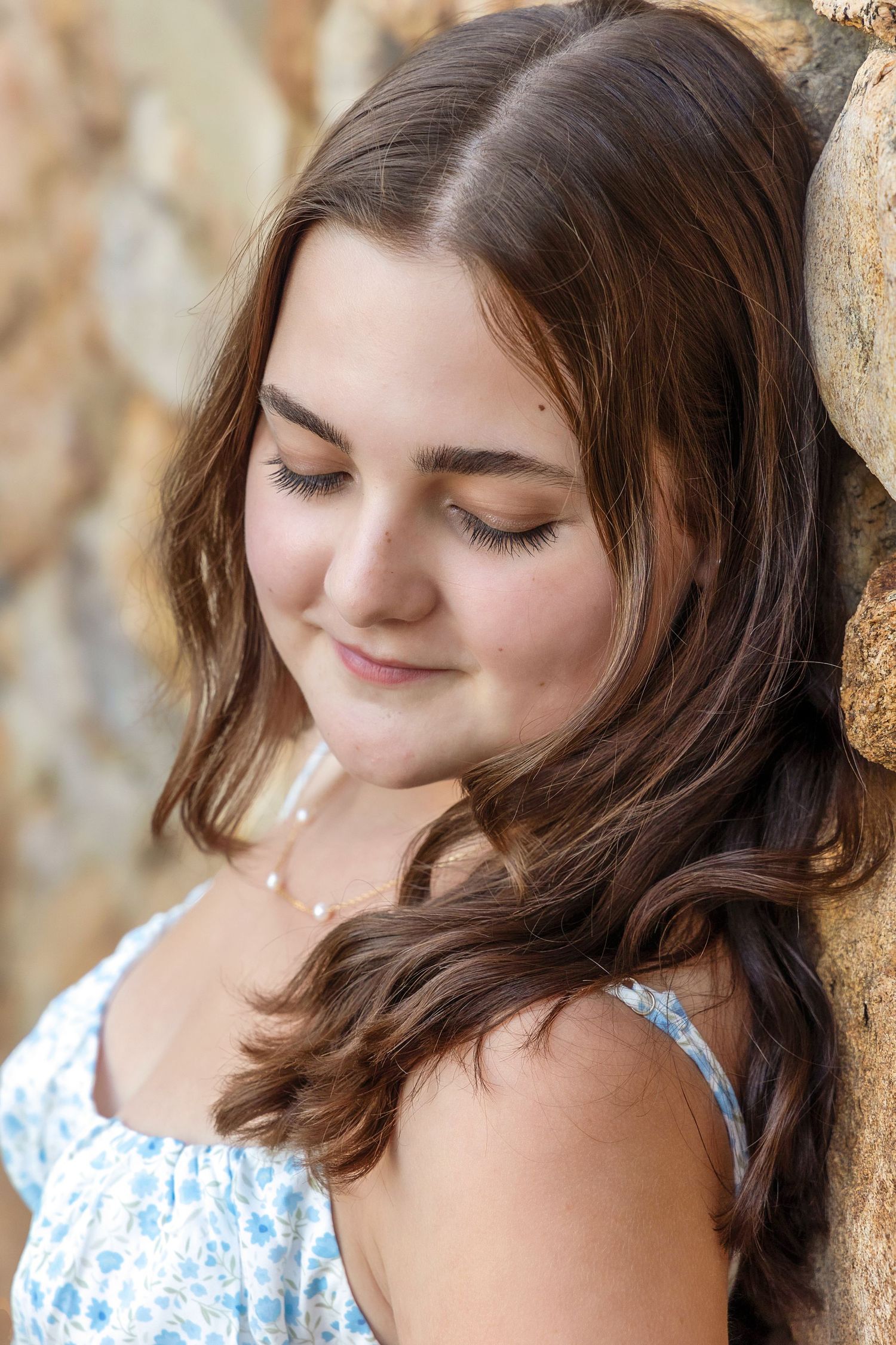 A woman in a white and blue floral dress leans against a textured wall with closed eyes and soft smile.