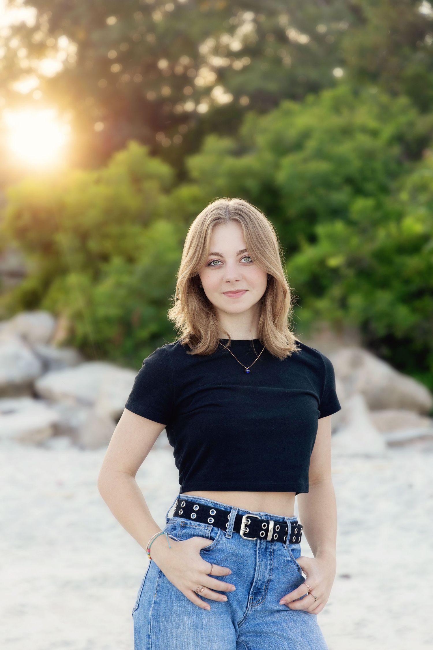 A person in a black crop top and high-waisted jeans poses outdoors at sunset with greenery in the background.
