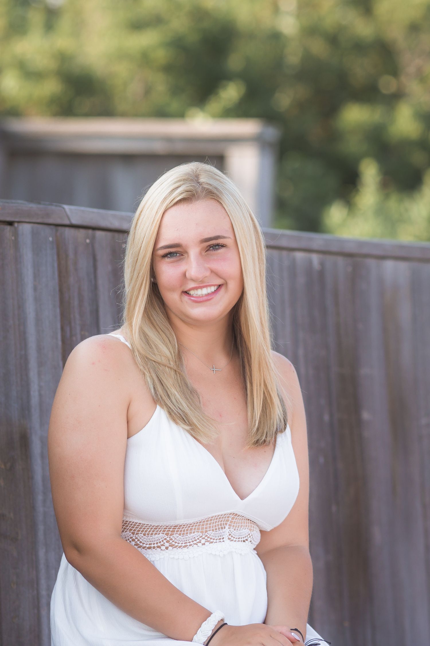 A person in a white sleeveless top poses outdoors against a wooden fence with greenery in the background.