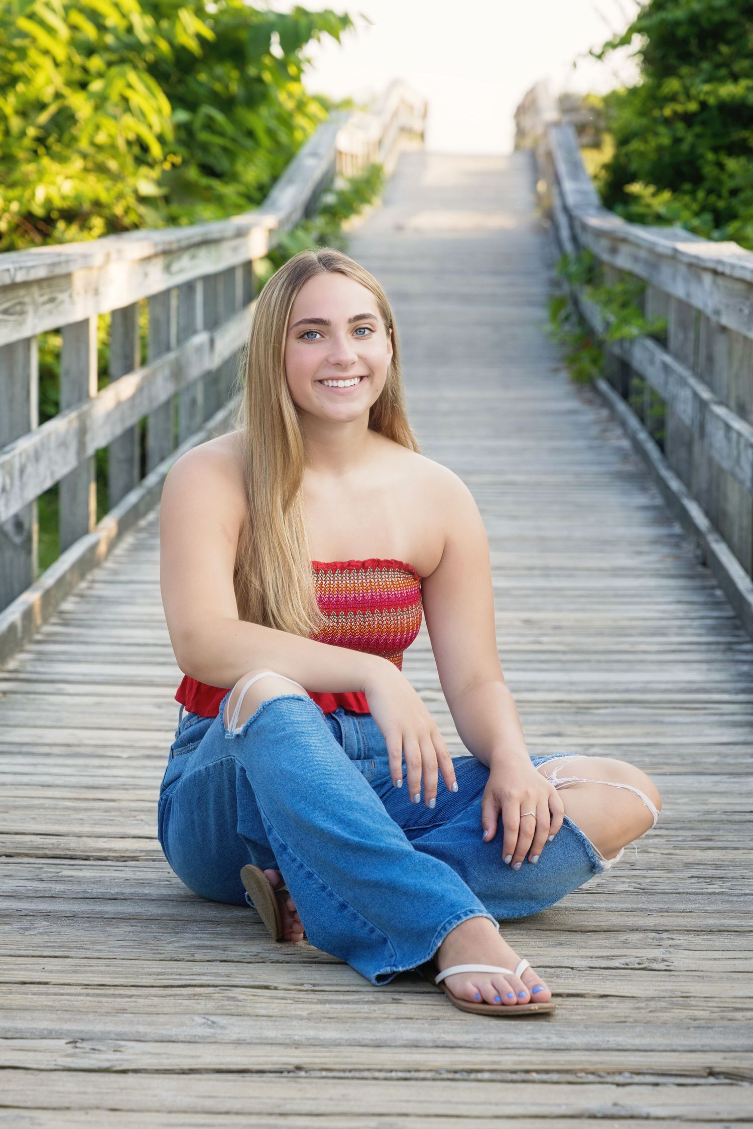 Person in red top and jeans sits casually on wooden boardwalk surrounded by lush summer greenery.