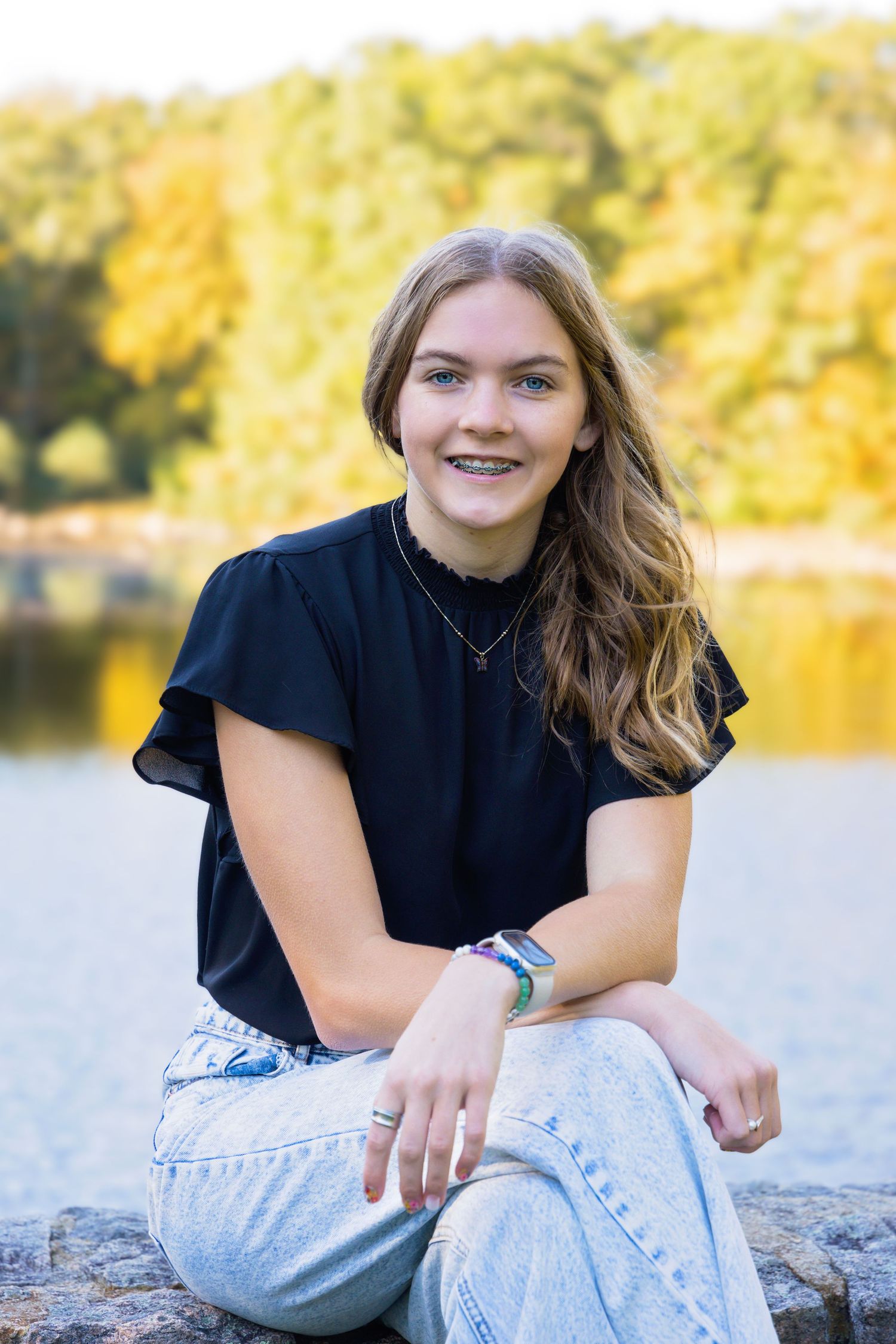 A person in a black shirt and light jeans sits casually on rocks by a lake with autumn foliage in the background.
