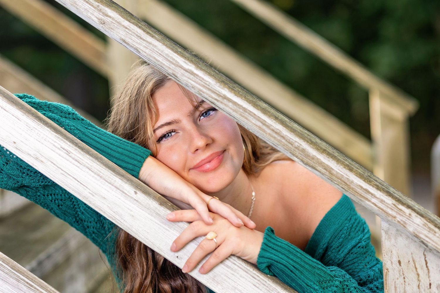Young woman in teal sweater smiling while resting head on hand near white window frame.