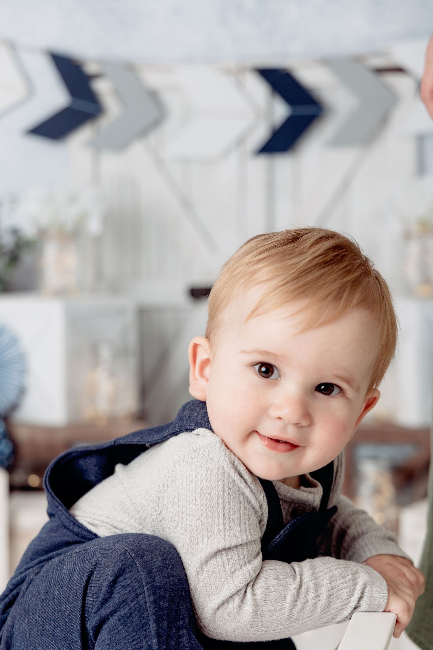 A baby wearing a gray sweater and navy pants smiles while lying on a white surface.