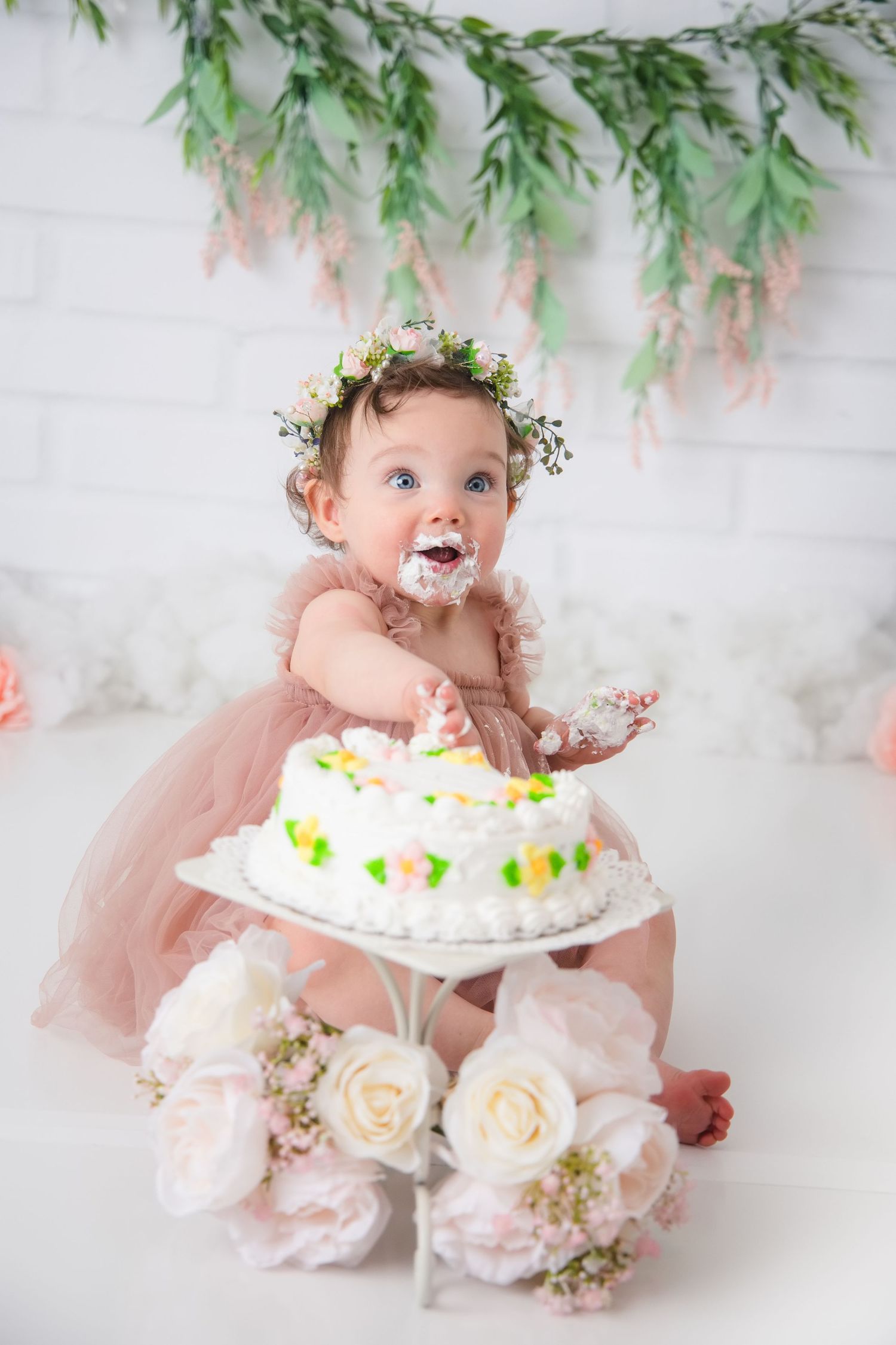 A baby wearing a floral crown enjoys smashing and tasting a white birthday cake during a cake smash photo session.