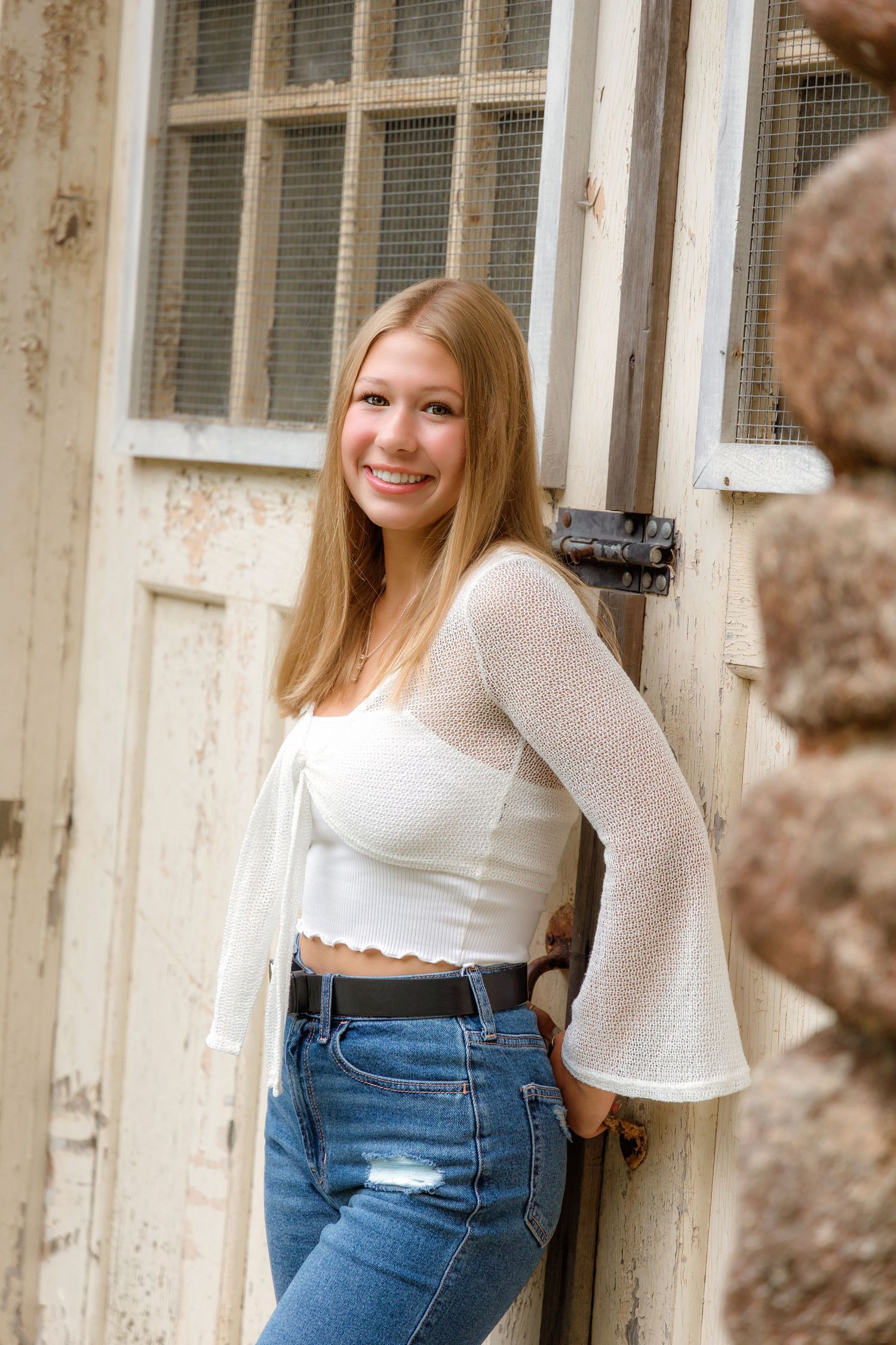 A person in a white bell-sleeve top and distressed jeans poses against a rustic door with mesh window panels.
