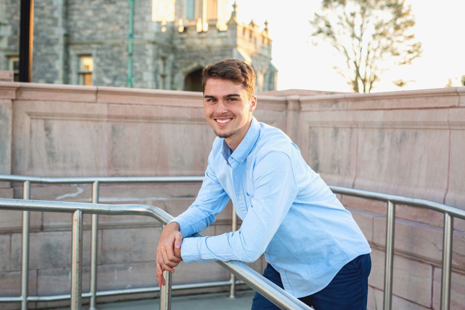 A person in a light blue sweater leans against a railing with a stone building in the background.