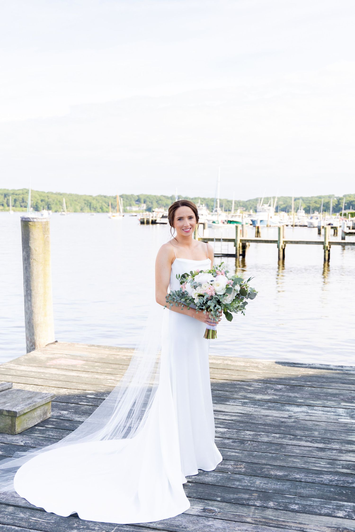 A bride in a flowing white wedding dress stands elegantly on a wooden dock overlooking a serene waterfront.