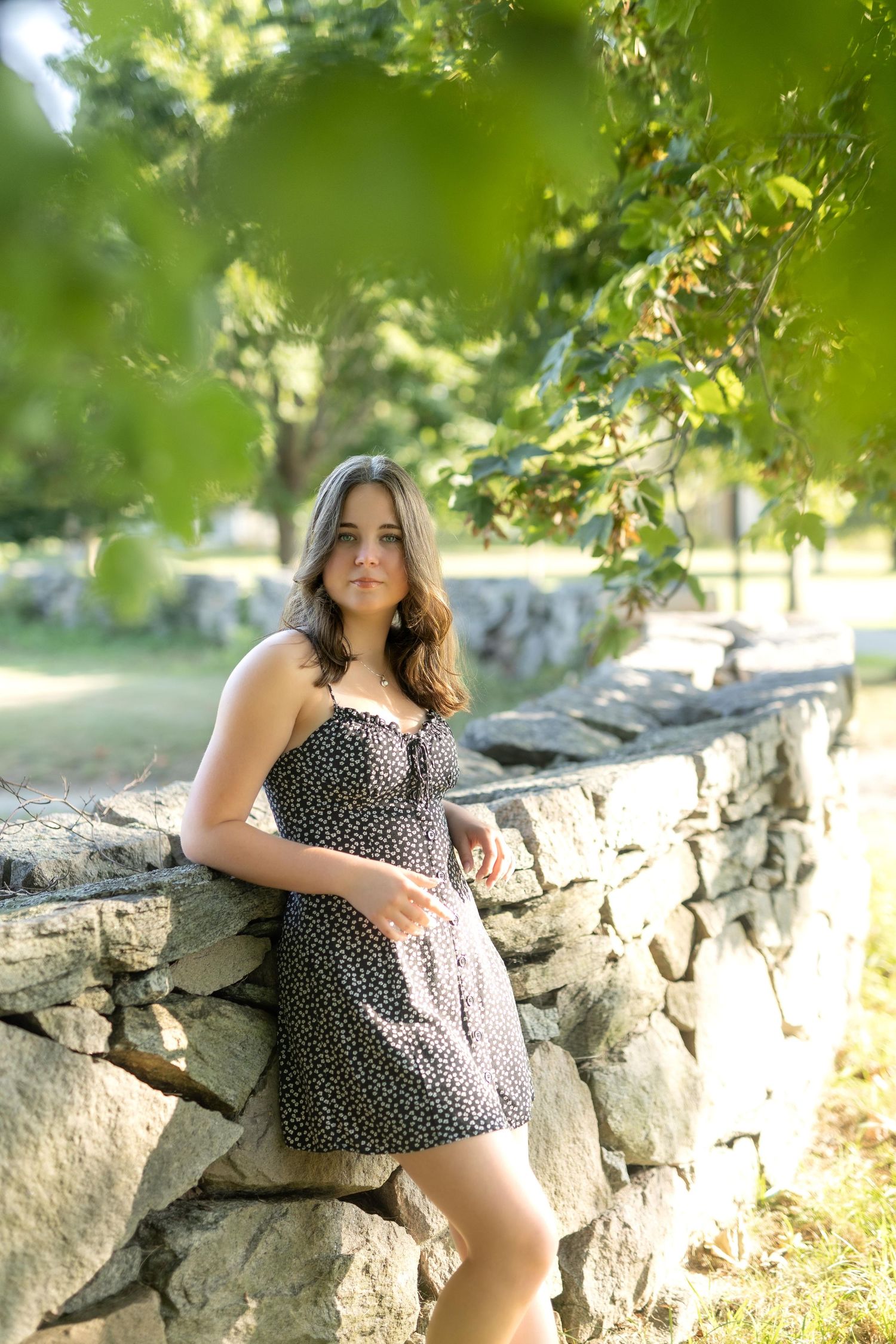 A person in a polka dot sundress leans against a rustic stone wall on a sunny summer day in a park setting.