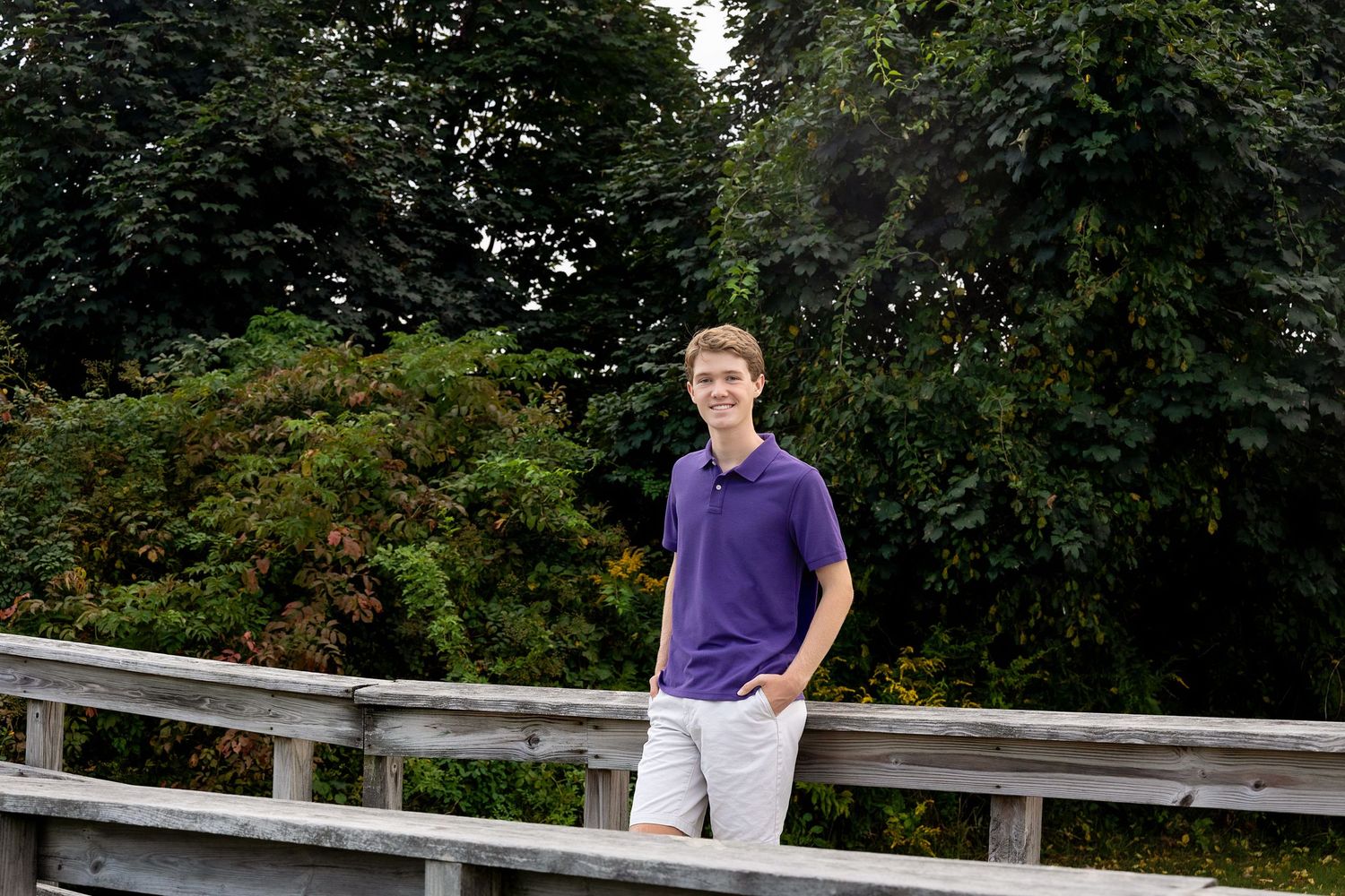 A person in a purple shirt stands on a wooden bridge railing in a lush green park setting.